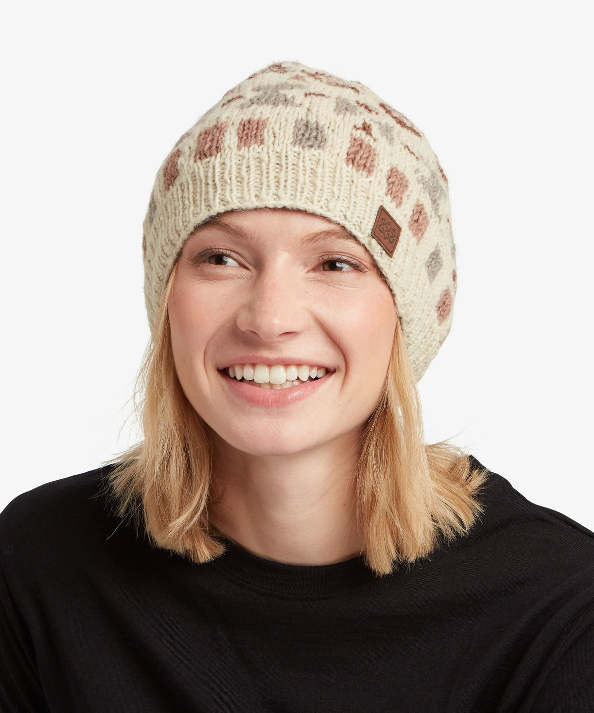 A smiling woman with blonde hair wearing a cream-colored knit beanie with a geometric pattern.