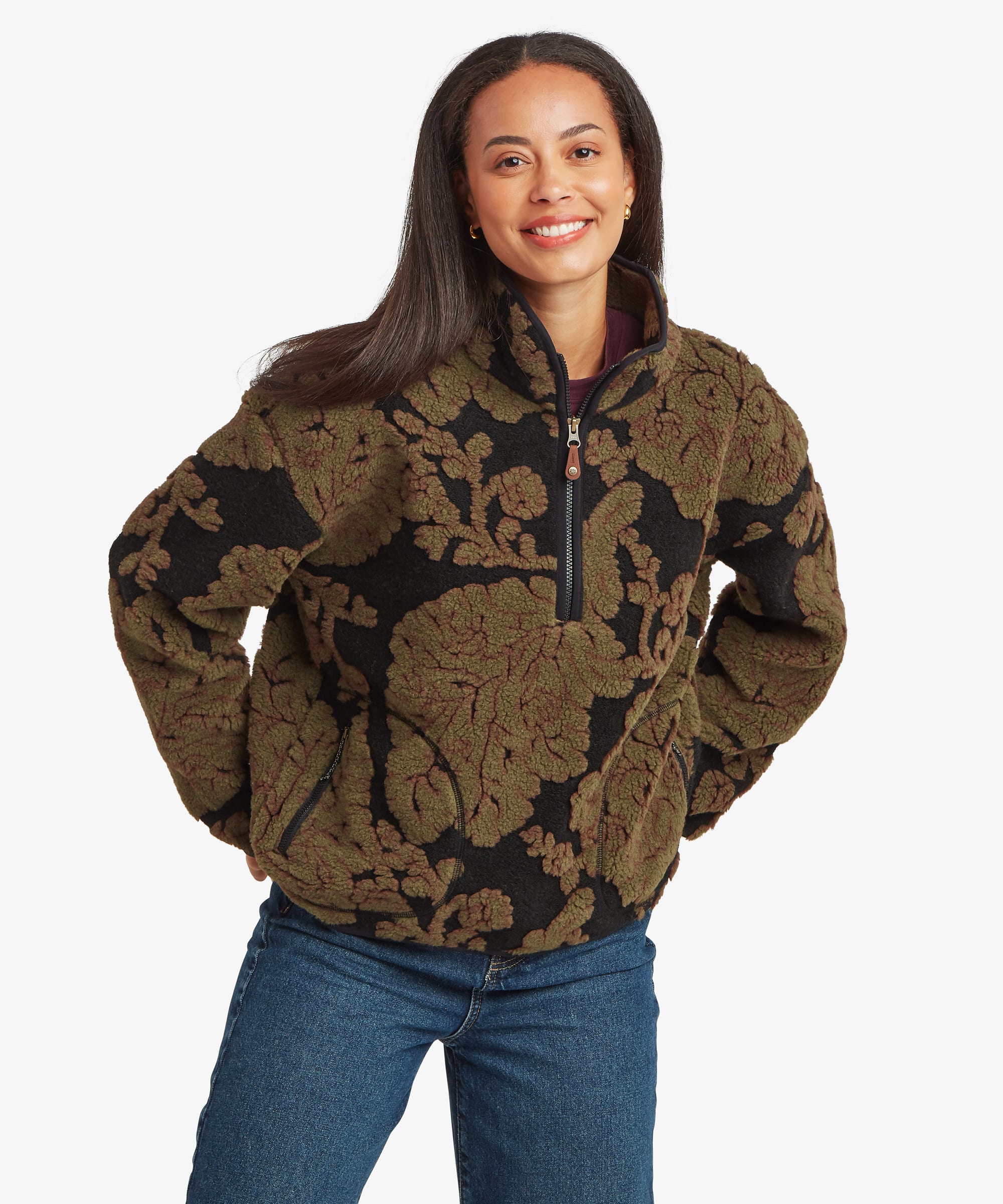 Woman standing facing forward in a studio setting wearing a dark green quarter-zip fleece pullover with an oversized black floral jacquard pattern. Paired with blue jeans, she stands confidently with her hands on her hips, highlighting the pullover's relaxed fit and high collar.