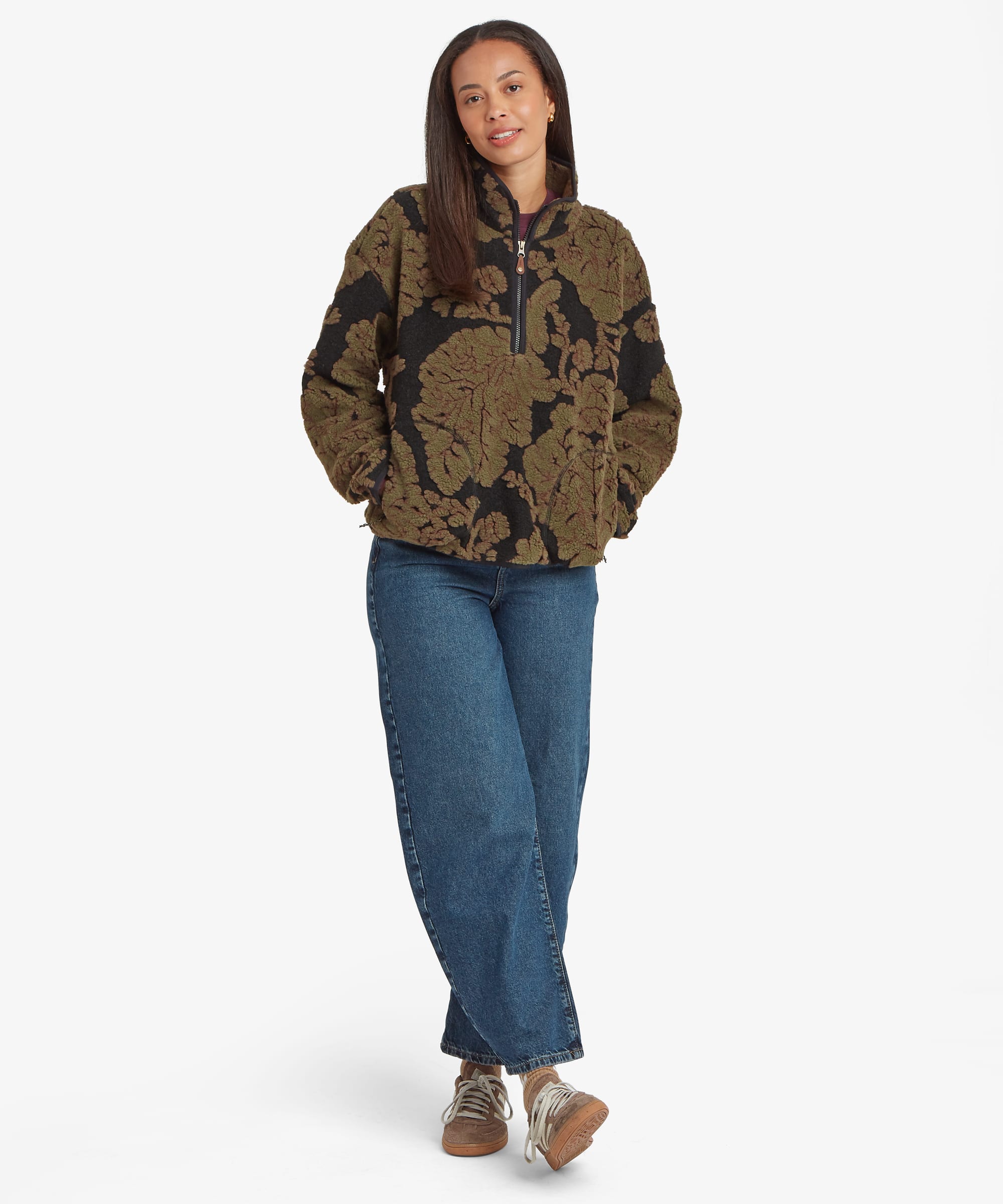 Full-length studio image of a woman wearing a dark green floral-patterned quarter-zip fleece pullover paired with blue jeans. The image showcases the garment's overall silhouette, stand collar, and bold botanical design.