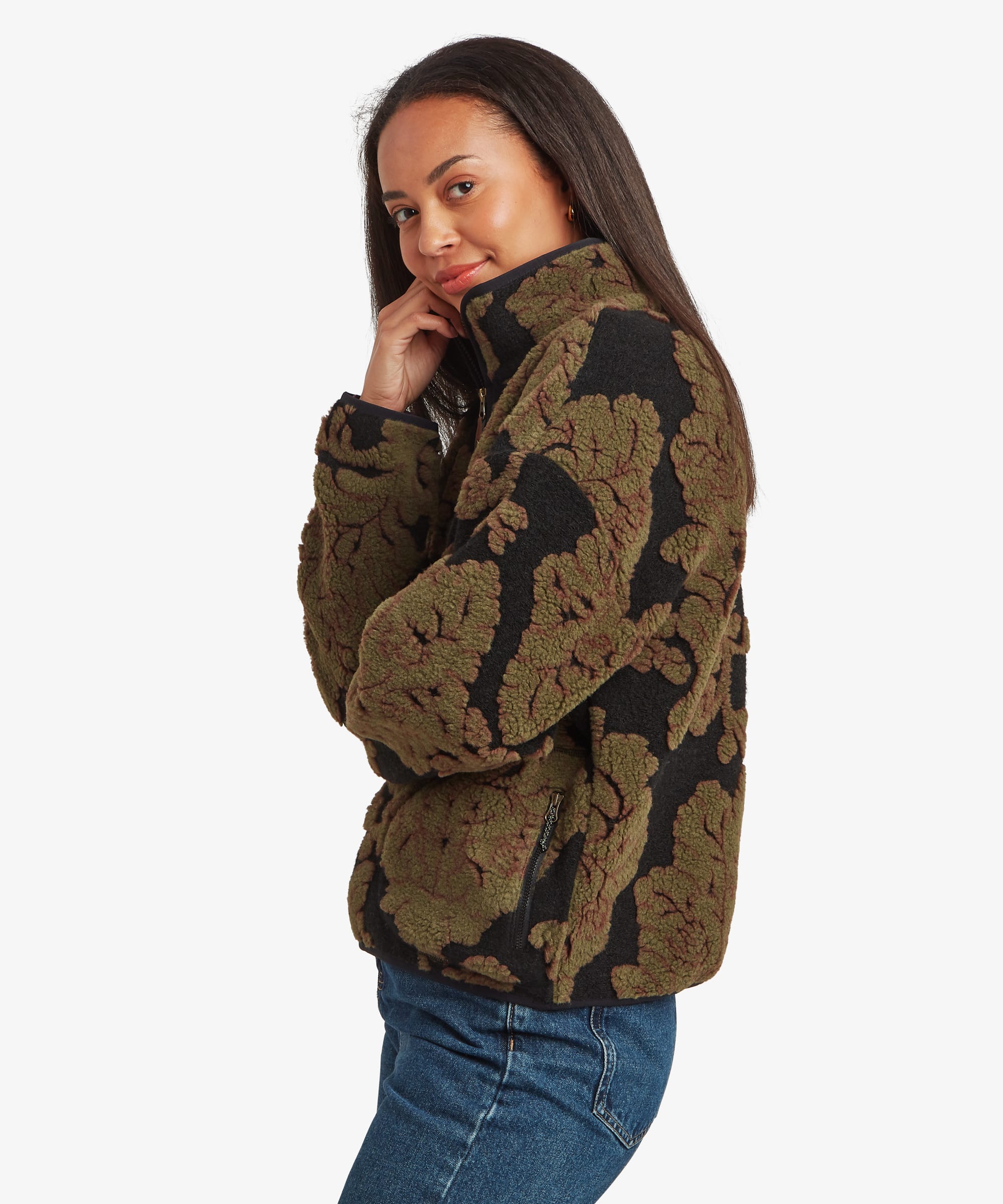 Side-profile studio view of a woman wearing a dark green quarter-zip fleece pullover with an oversized floral jacquard pattern in black. Paired with blue jeans, she glances over her shoulder, showcasing the pullover's relaxed fit and high collar.