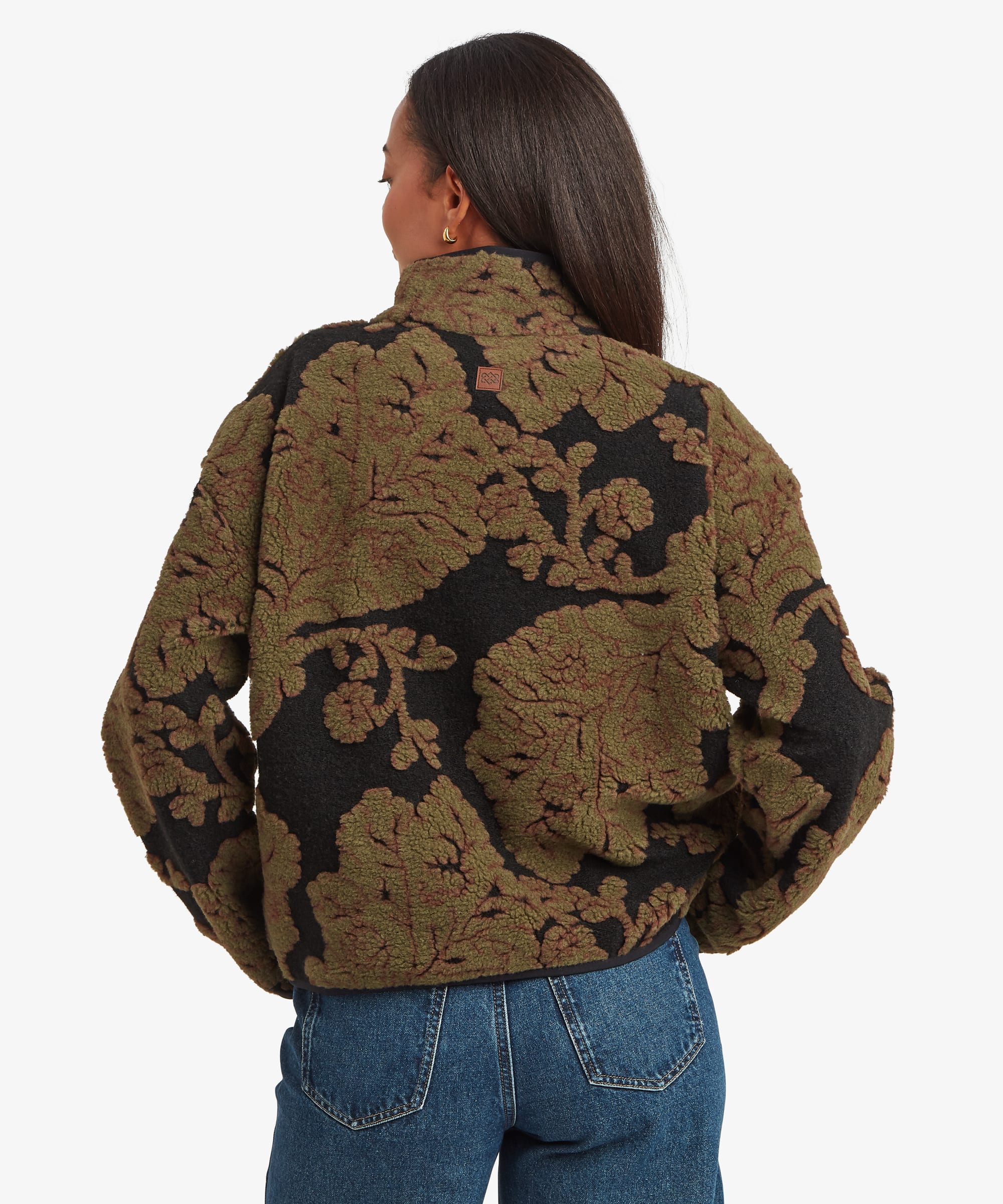 Rear studio view of a woman wearing a dark green quarter-zip fleece pullover with a large floral pattern across the fabric. Styled with blue jeans, the image highlights the back design and relaxed silhouette.