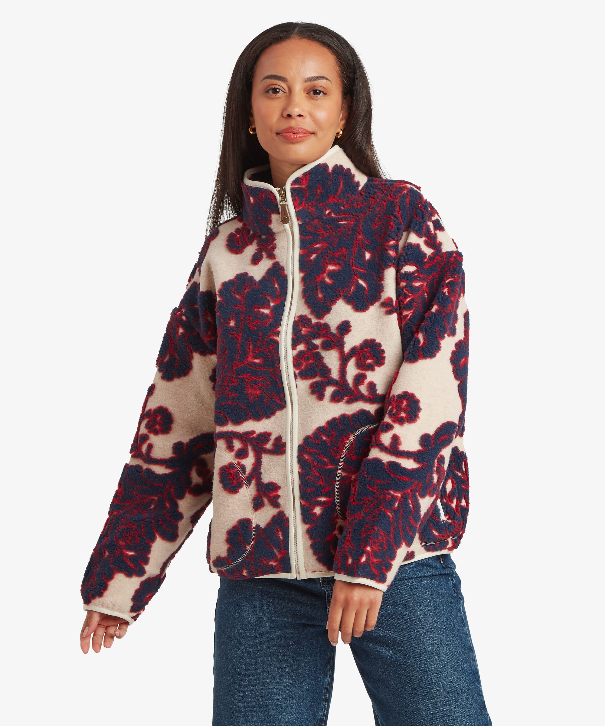 Woman standing facing forward in a studio setting wearing a cream full-zip fleece jacket with a bold burgundy floral pattern throughout. Paired with blue jeans, she walks naturally toward the camera, showcasing the jacket's front design and relaxed fit.