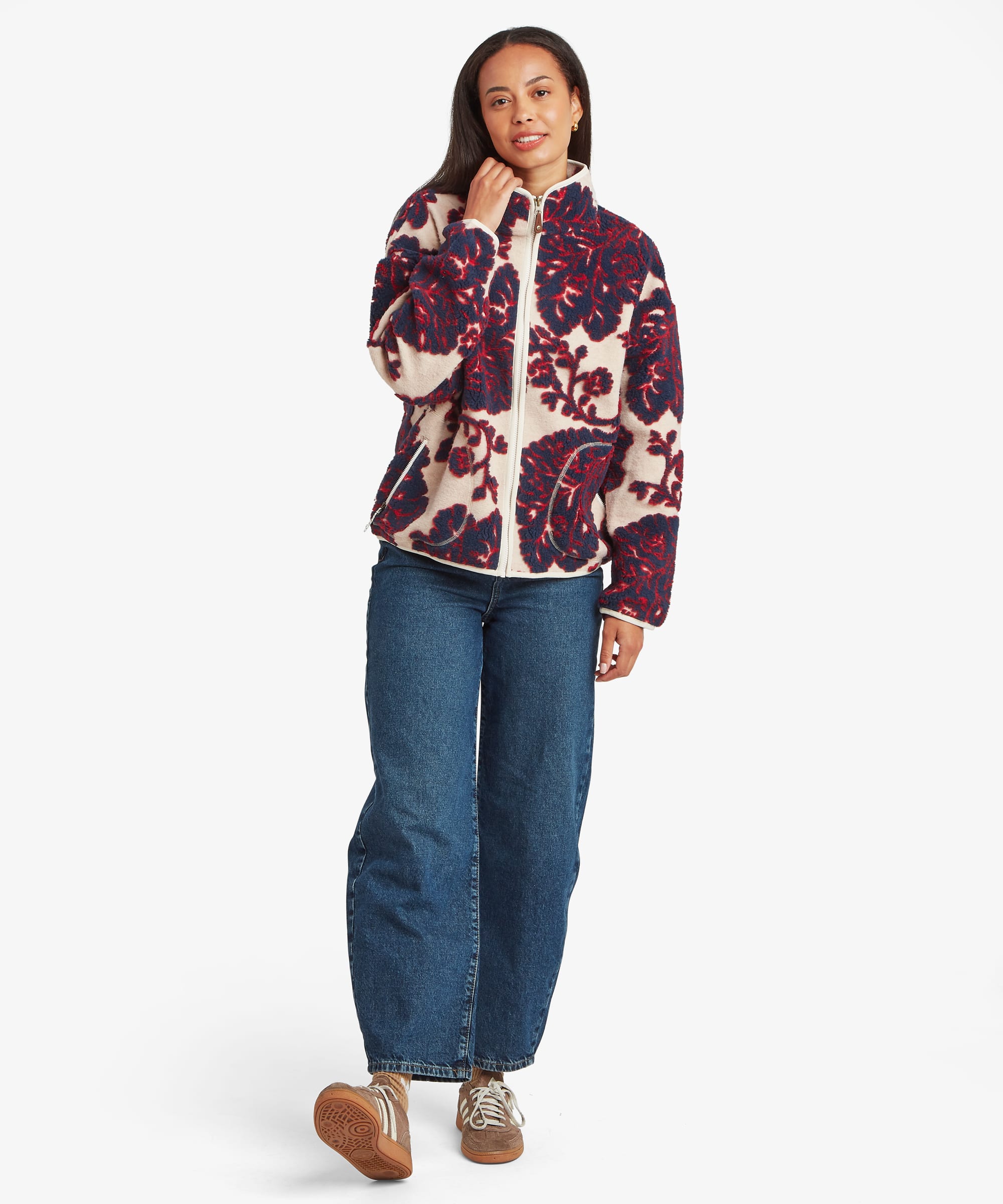Full-length studio image of a woman wearing a cream fleece jacket featuring a large burgundy floral print. Styled with blue jeans, the image highlights the jacket's overall silhouette, stand collar, and full-length zip closure.