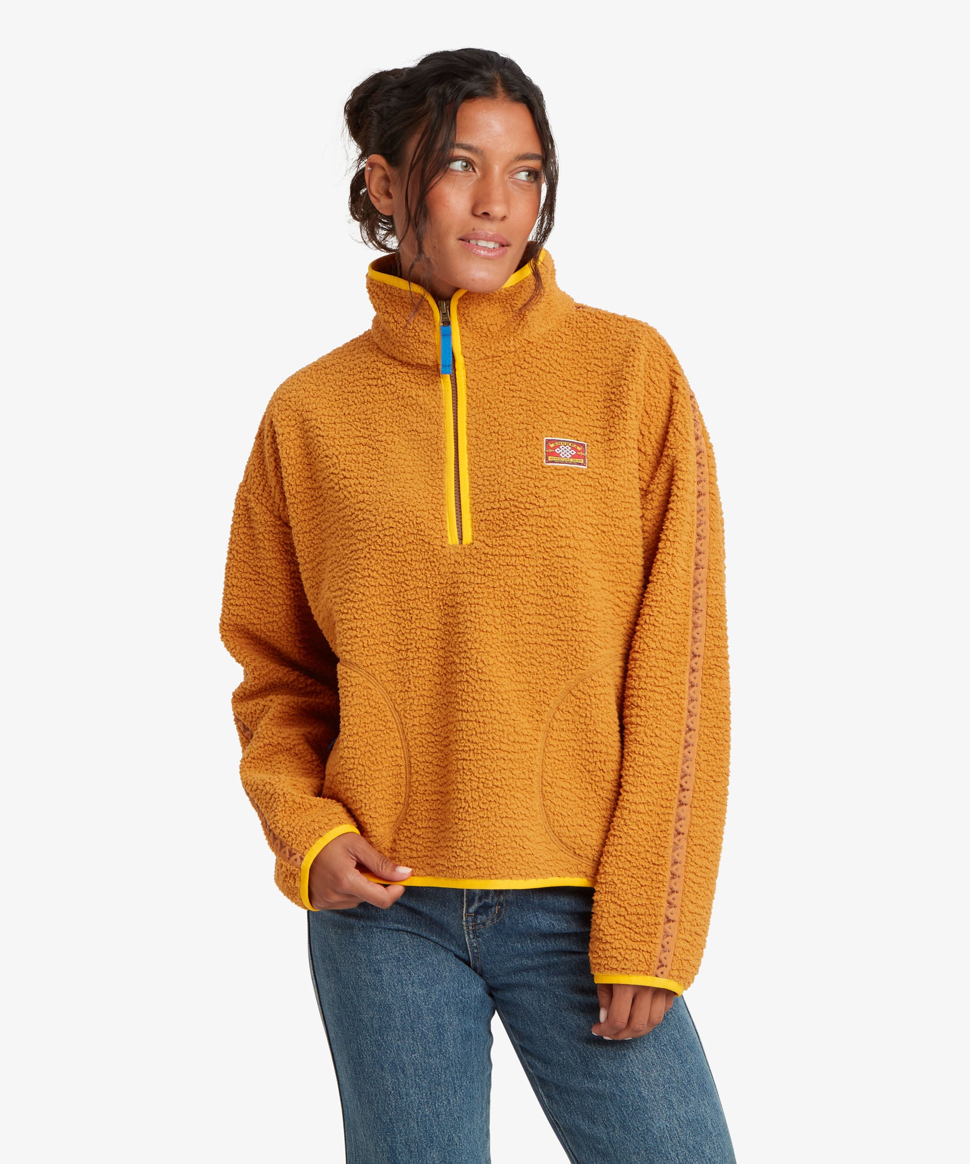 A woman wearing a textured golden-yellow quarter-zip fleece pullover with a small red and white logo patch on the chest, paired with blue jeans, against a light gray background.