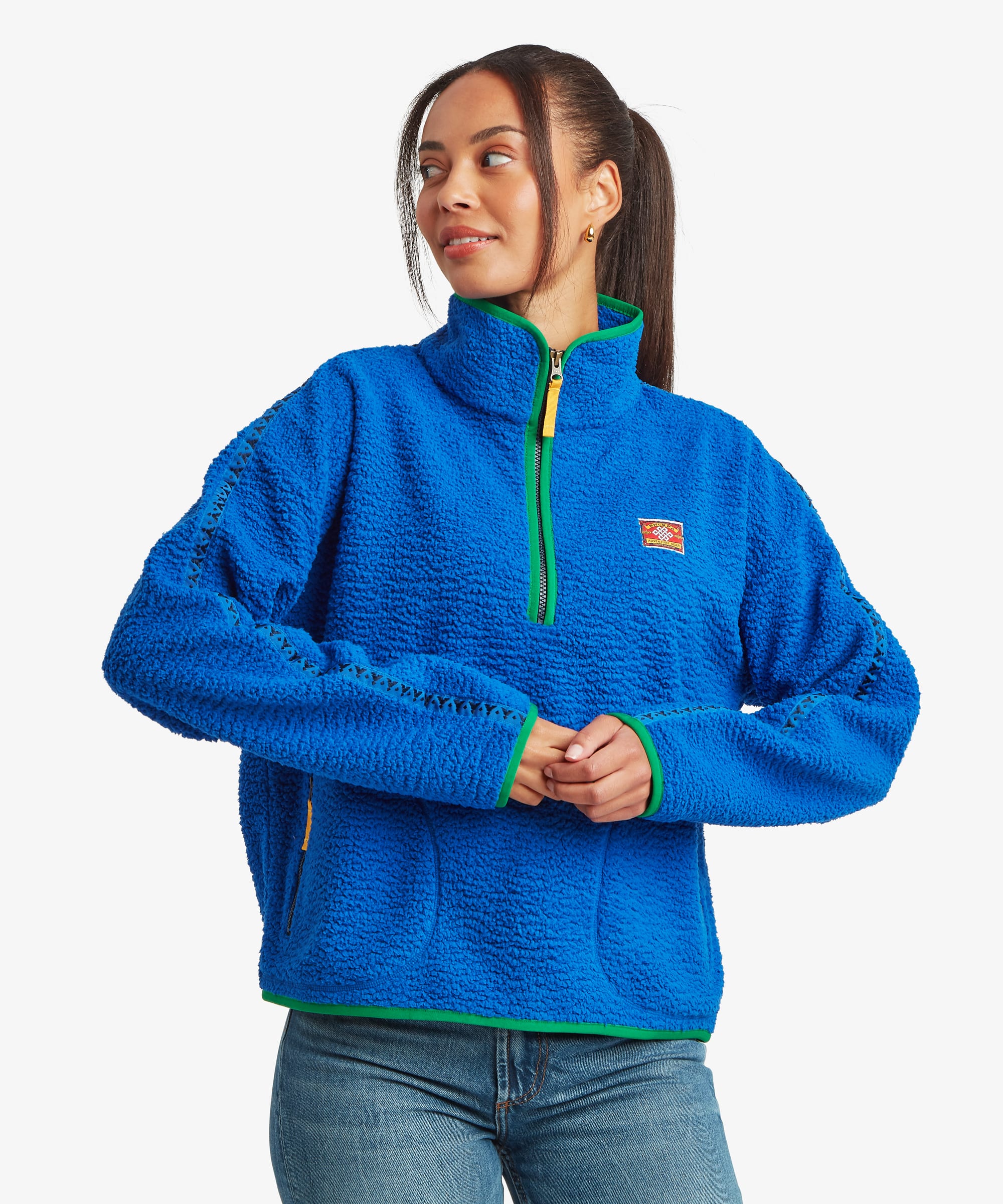 Woman standing against a plain studio background wearing a bright blue quarter-zip fleece pullover with contrasting green trim at the collar and zipper. Paired with blue jeans, she lightly clasps her hands while showcasing the front fit of the garment.