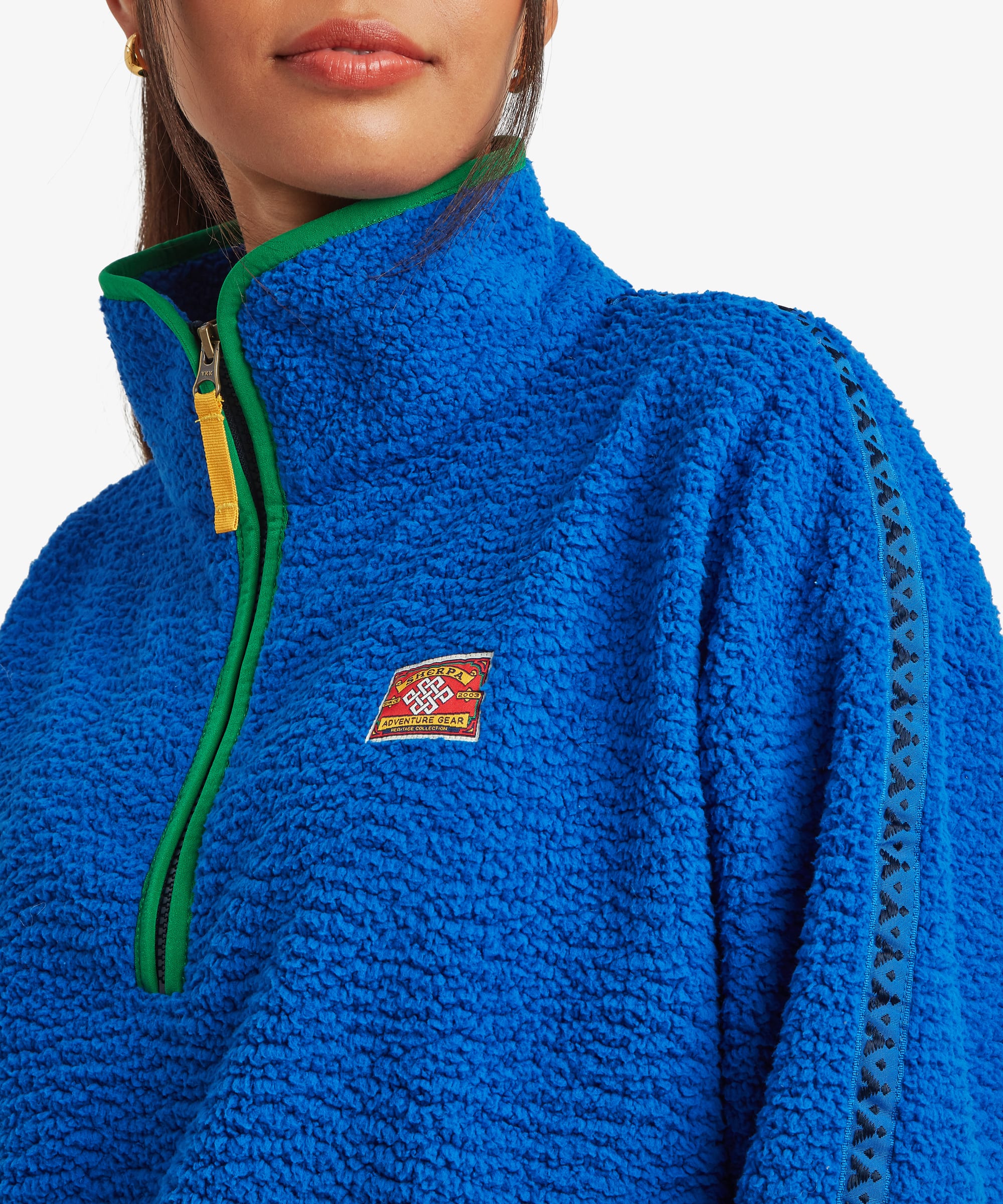 Close-up detail of a bright blue fleece pullover showing the quarter-zip opening, high collar, chest logo patch, and contrasting green trim around the zipper.
