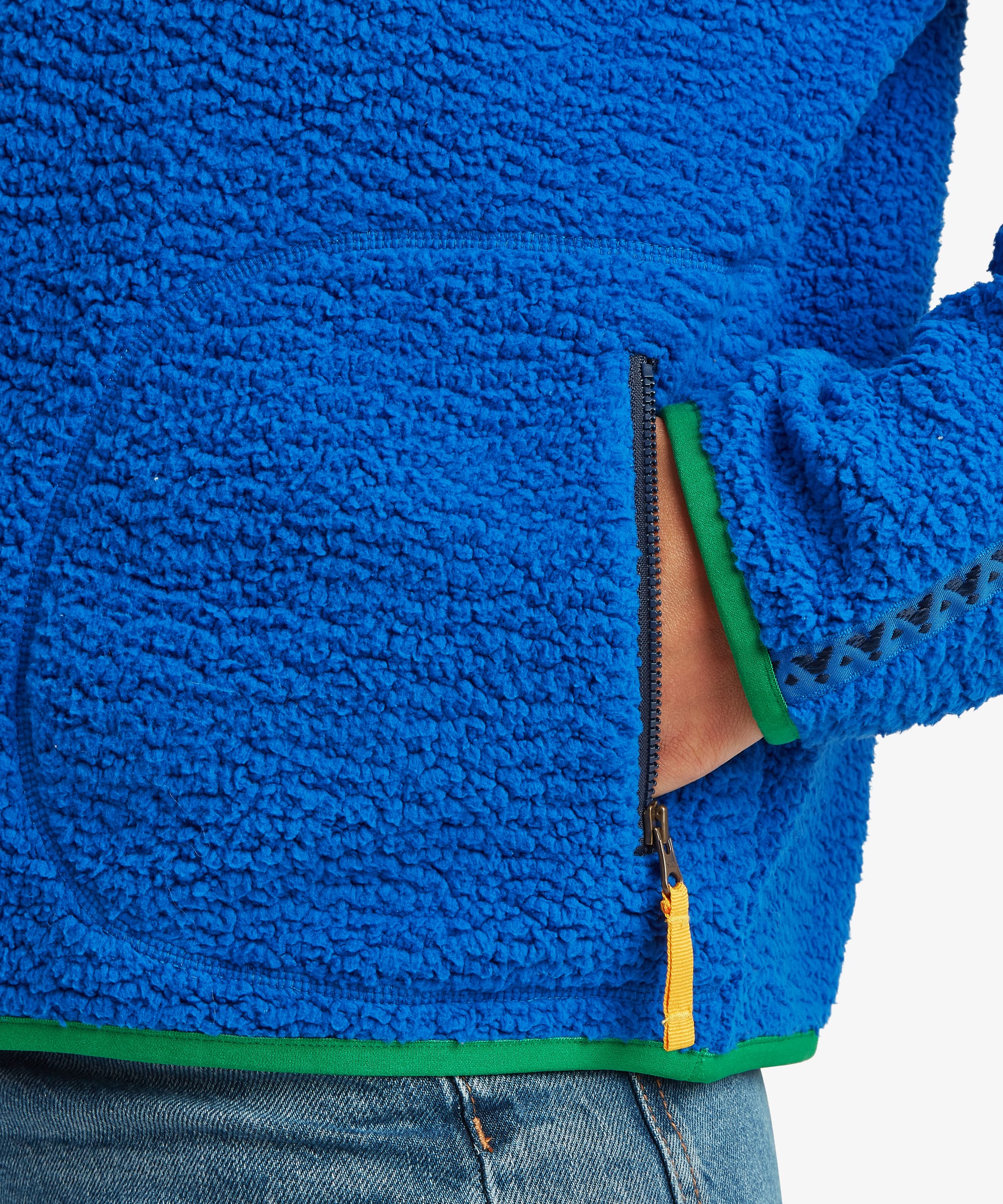 Detailed close-up of the bright blue fleece pullover highlighting the textured fabric, zippered pocket opening, and contrasting green trim along the hem and pocket edge.