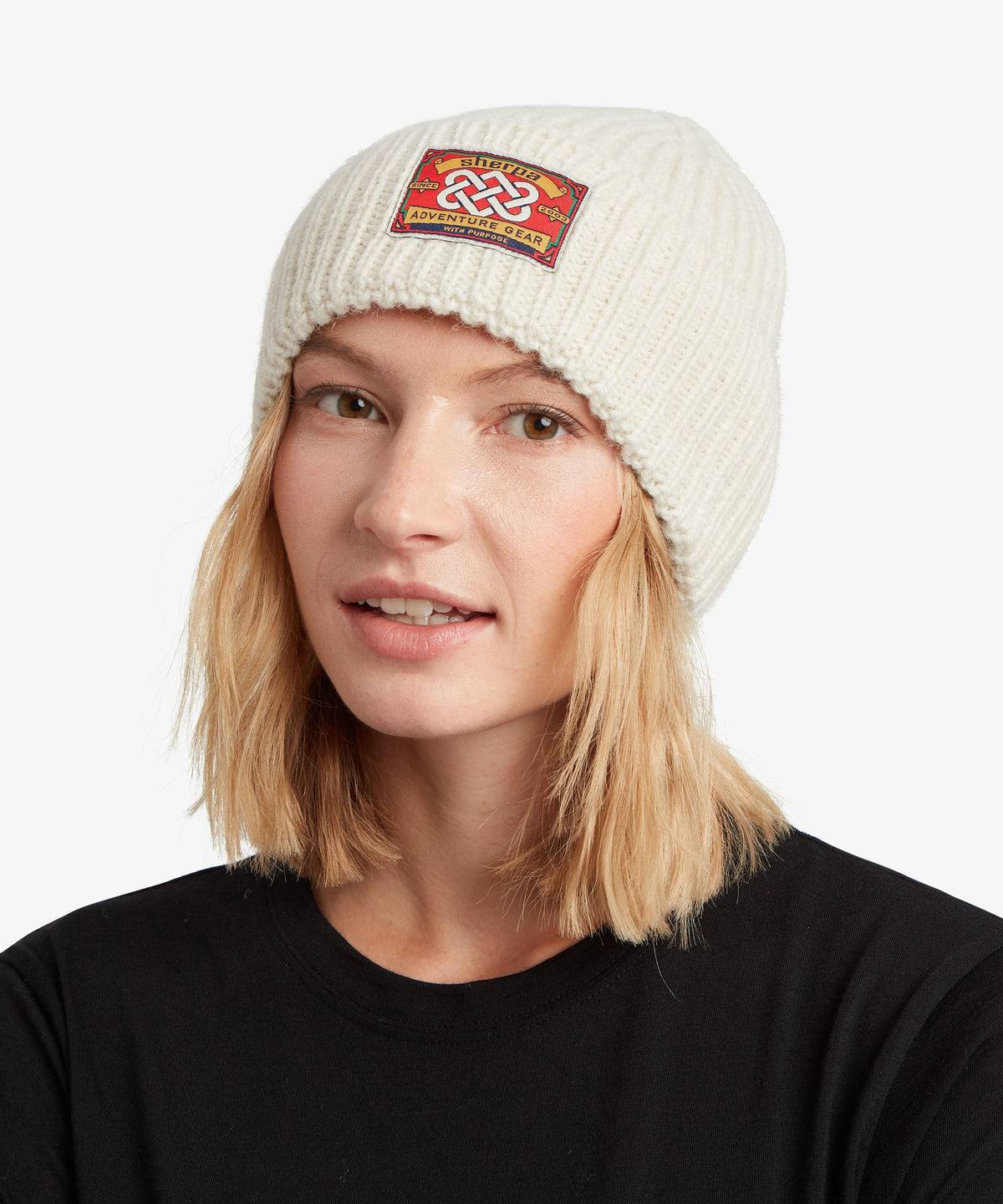 A woman wearing a cream-colored ribbed knit beanie with a colorful embroidered patch on the front.