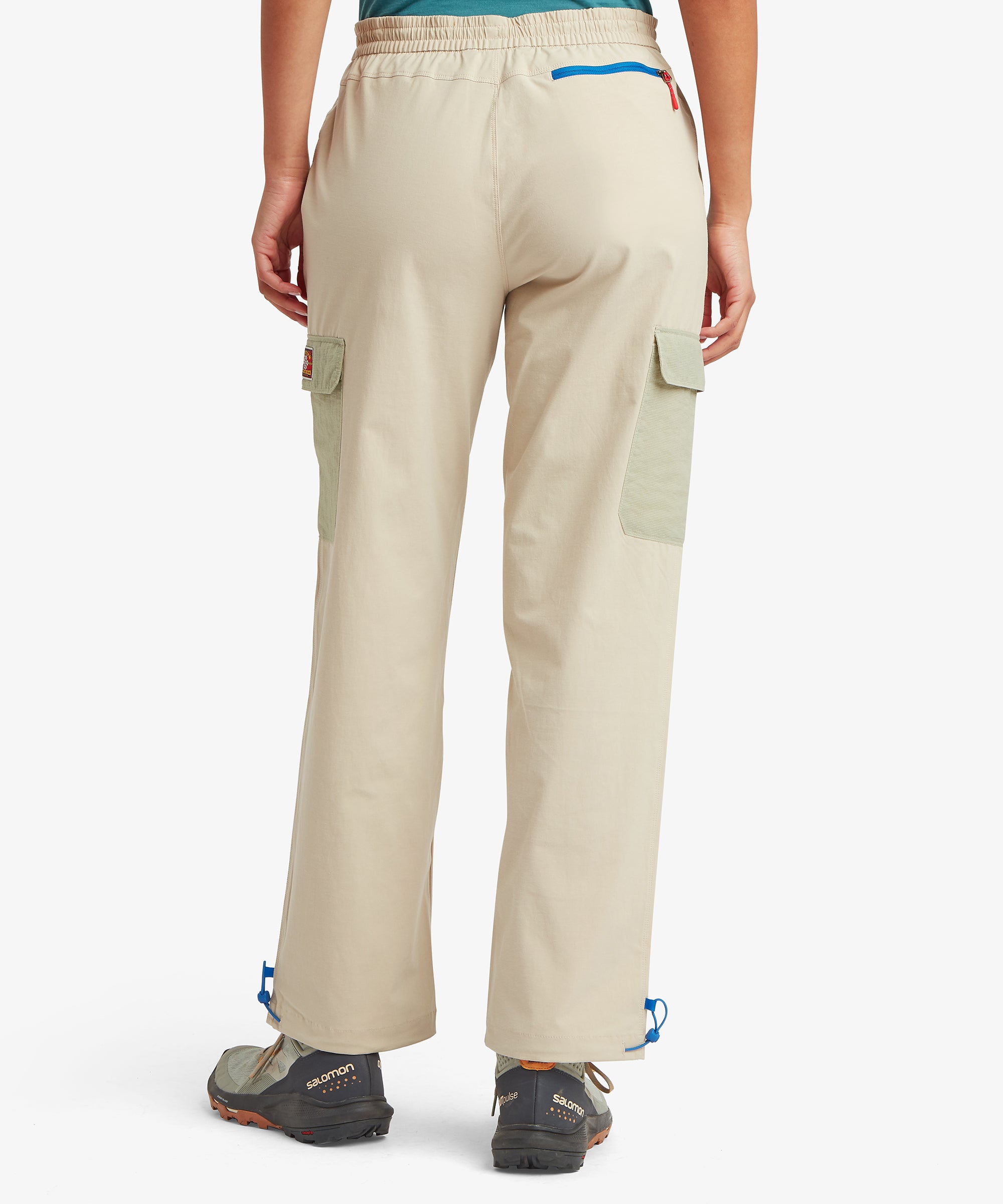 Back view of beige adventure trousers with dual side cargo pockets, a blue‑zip security pocket at the waistband, and straight‑leg styling suited for outdoor activities.