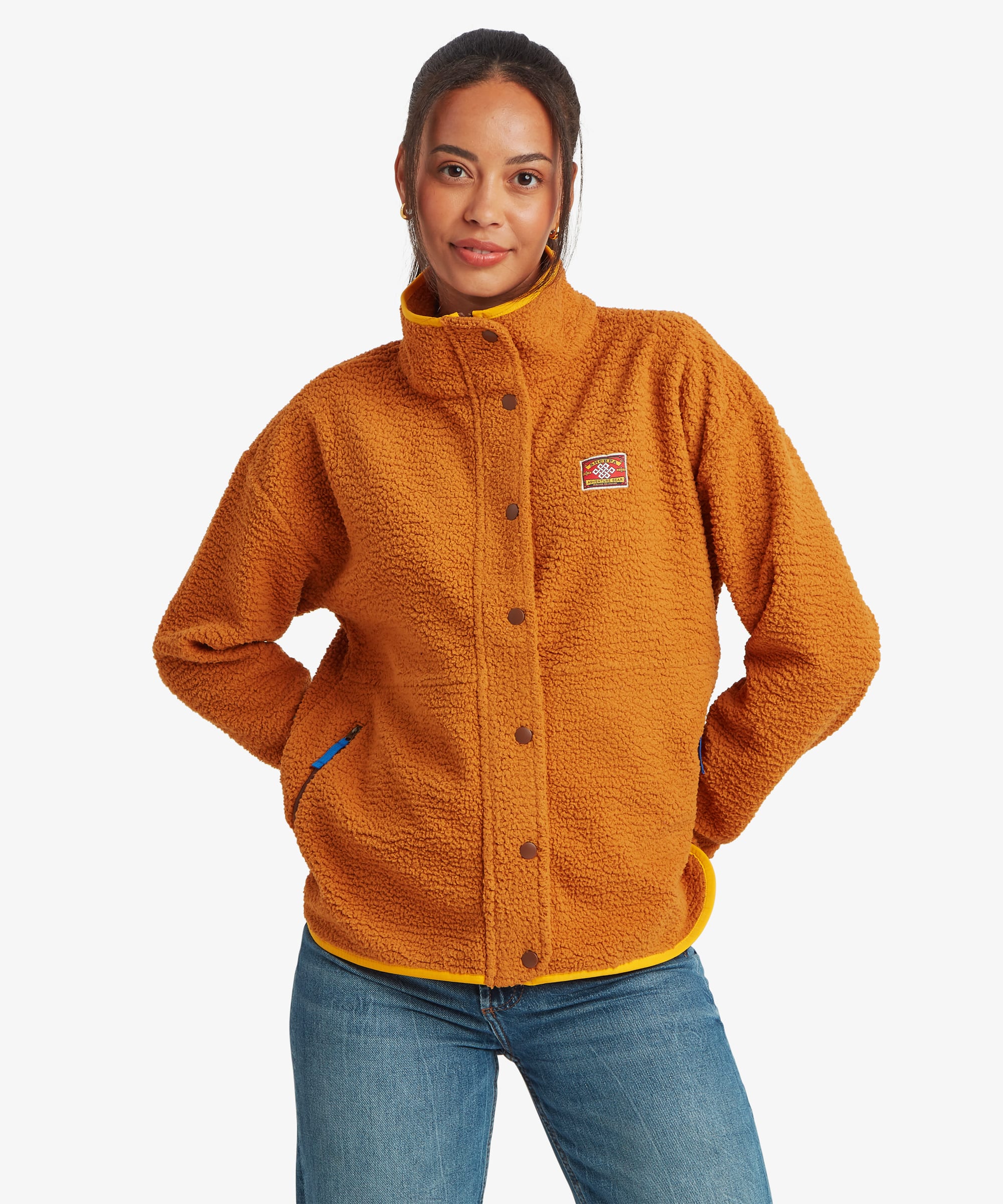 Woman standing facing forward in a studio setting wearing a mustard-yellow full-zip fleece jacket with a high stand collar and contrasting trim details. Paired with blue jeans, she rests one hand in her pocket while showcasing the jacket's relaxed fit.