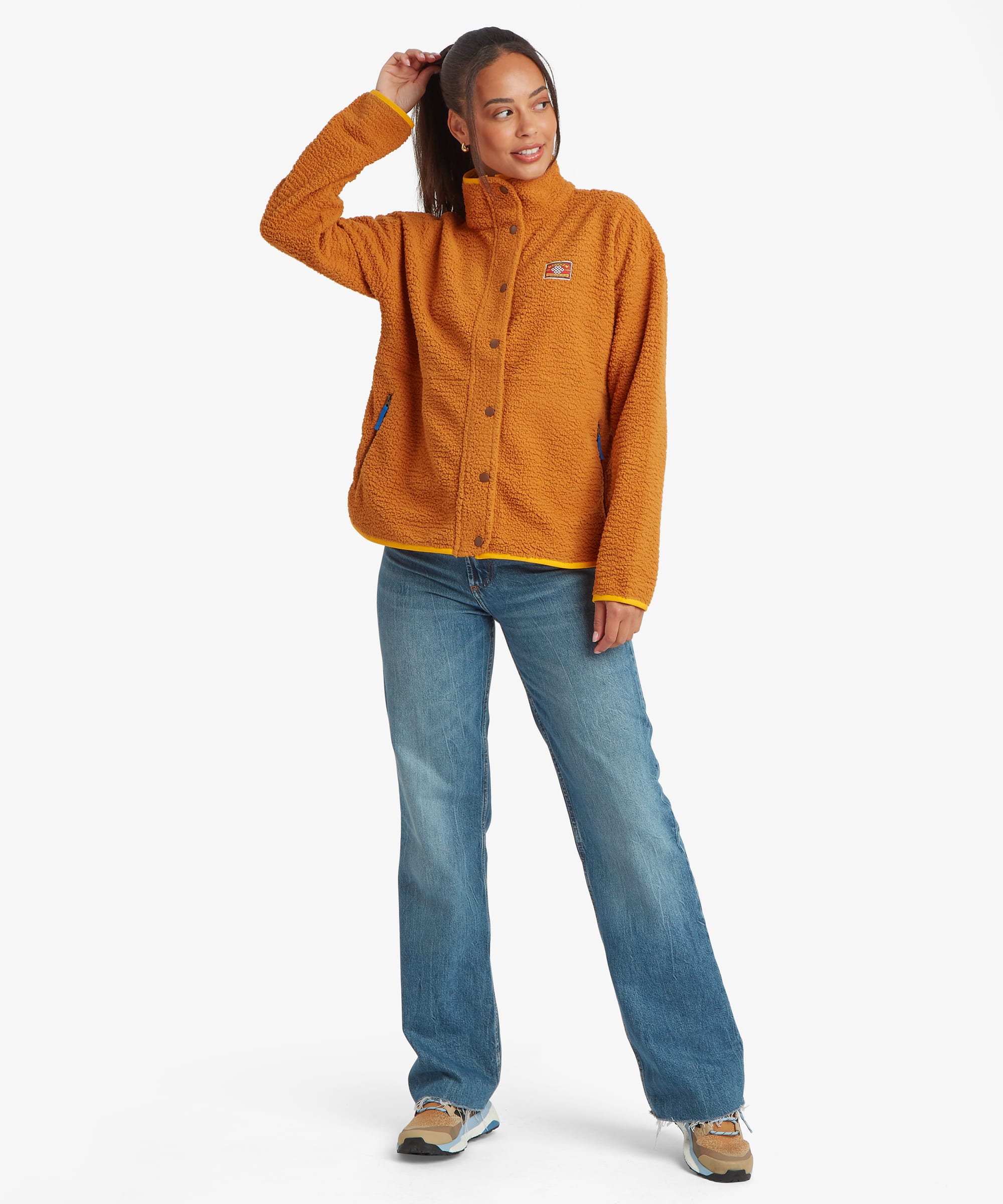 Full-length studio image of a woman wearing a mustard-yellow fleece jacket with a full zip front and high collar. Styled with blue jeans, she stands casually with one hand raised, highlighting the jacket's overall silhouette and length.