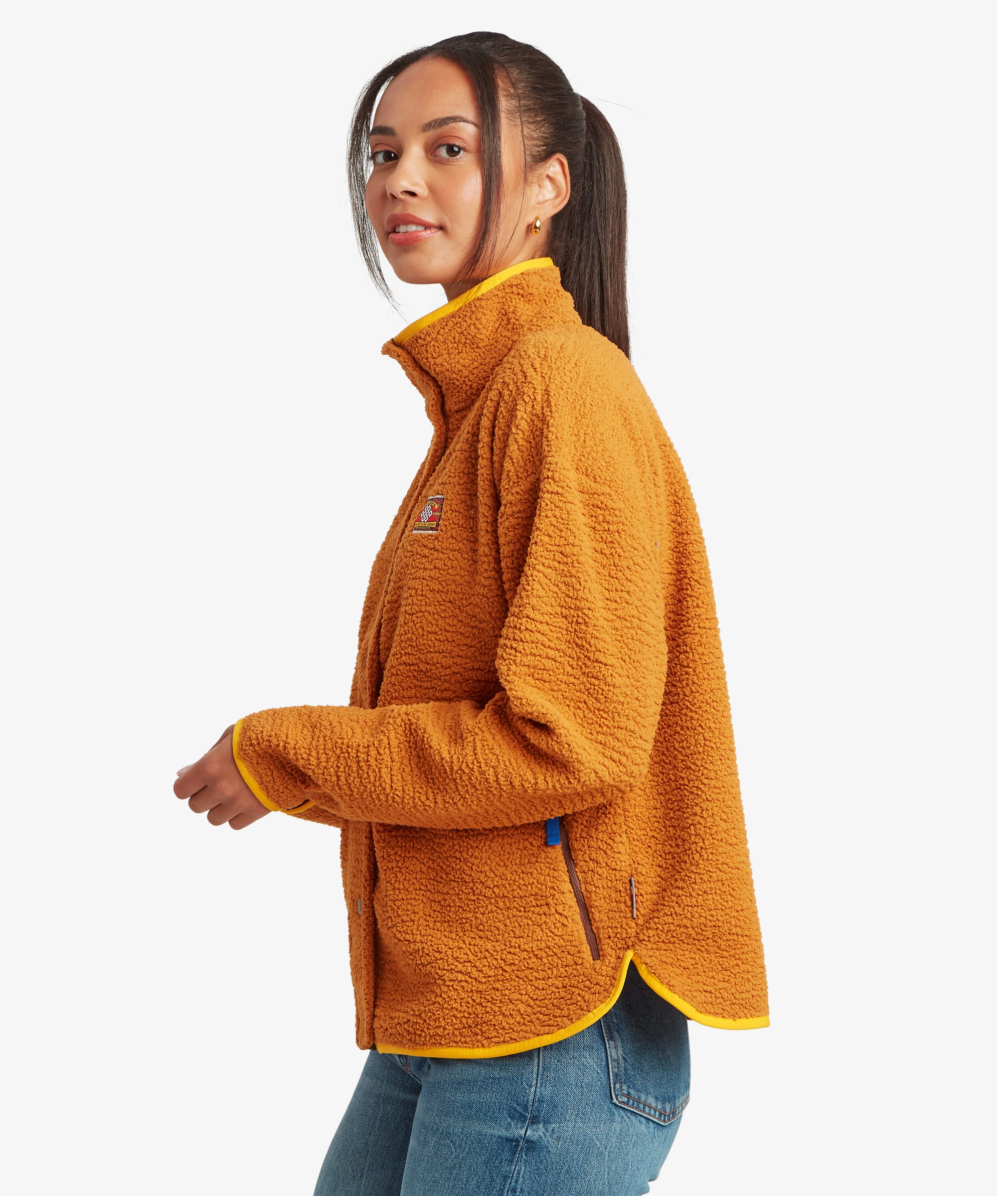 Side-profile studio view of a woman wearing a mustard-yellow full-zip fleece jacket with a high collar and relaxed fit. The image showcases the shape of the jacket through the body and sleeves.