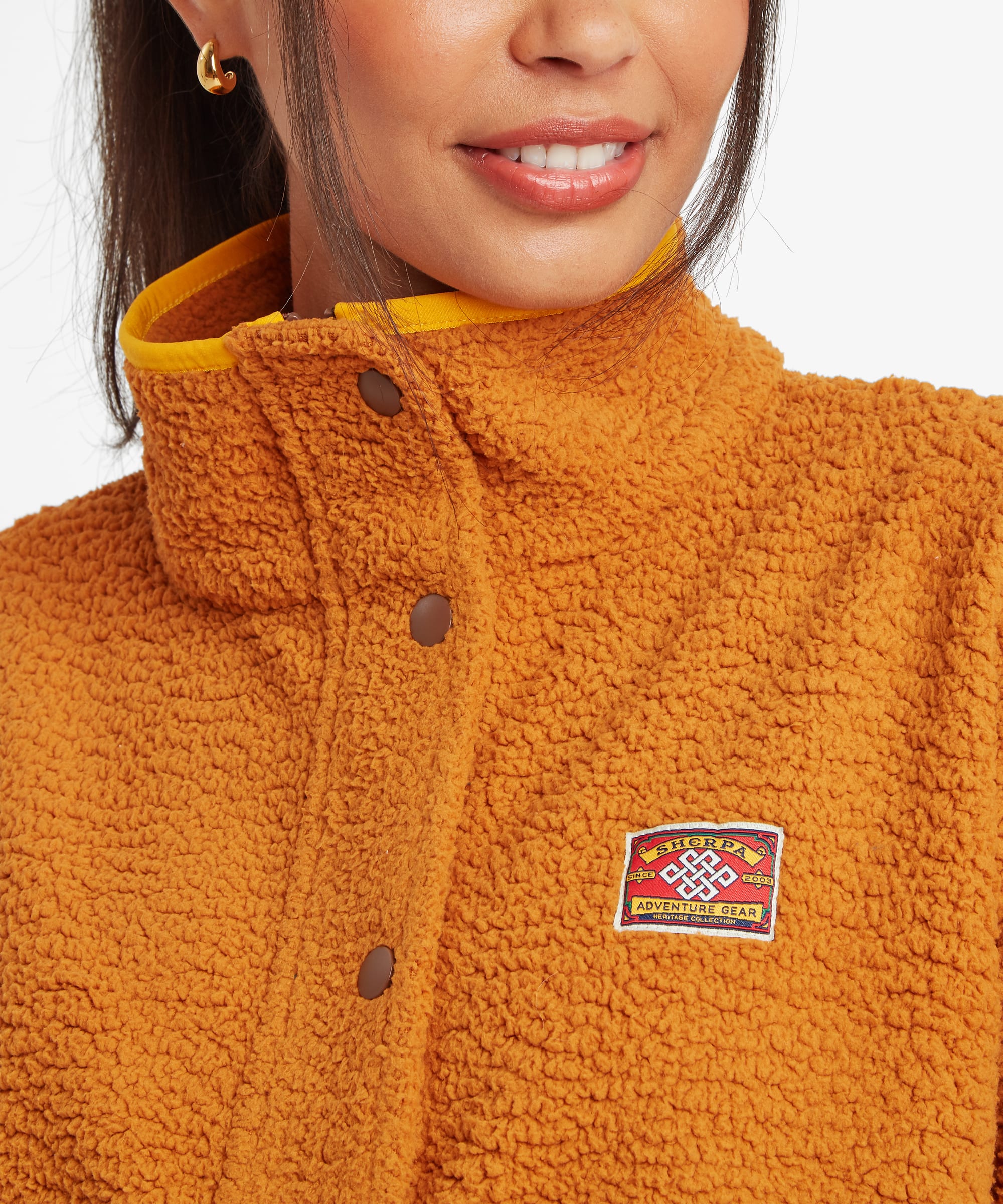 Close-up detail of a mustard-yellow fleece jacket showing the high stand collar, snap-button placket, chest logo patch, and textured fleece fabric.