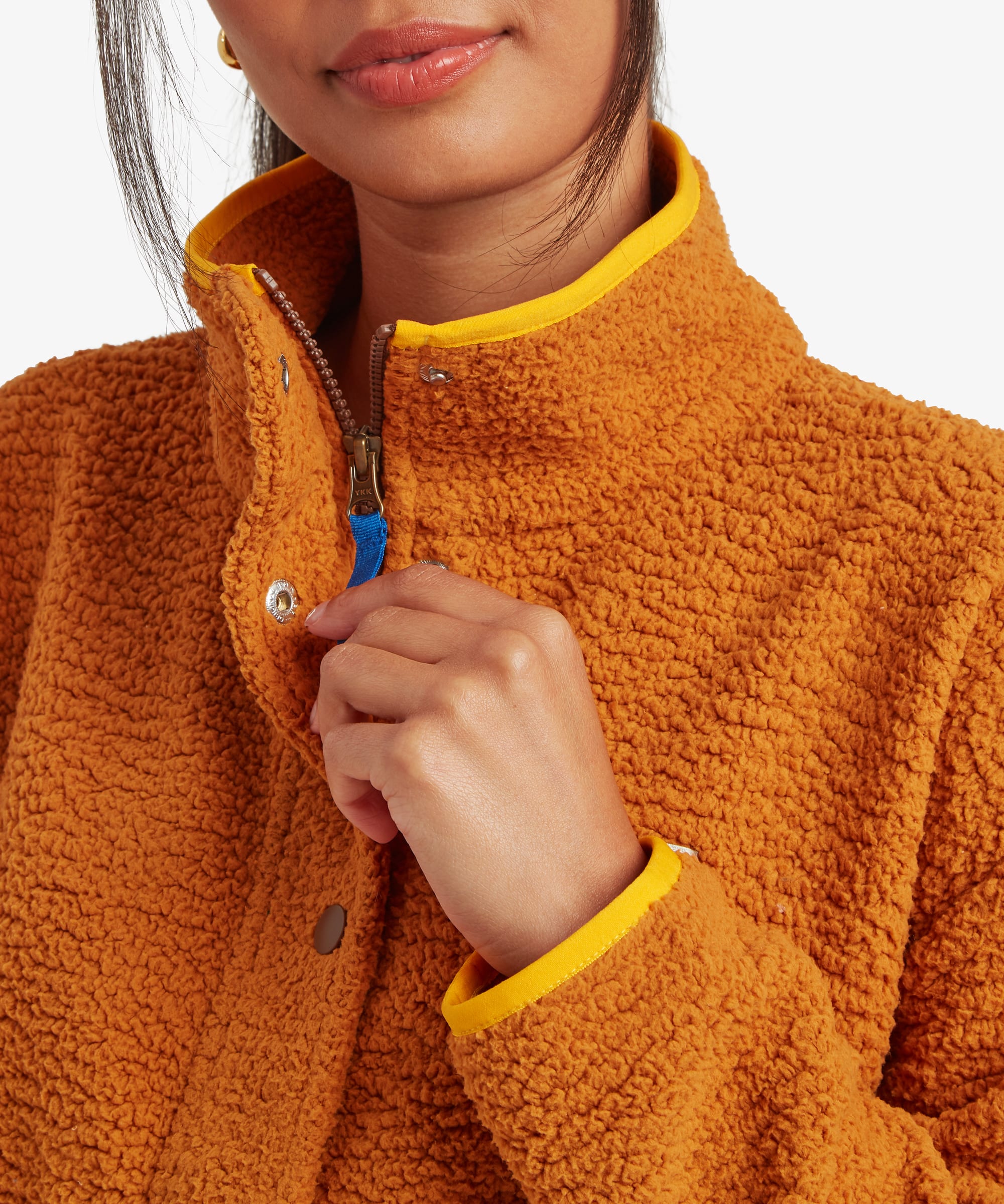 Close-up image of a model adjusting the collar of a mustard-yellow fleece jacket. The image highlights the high neckline, contrasting trim, snap closure details, and soft textured fabric.