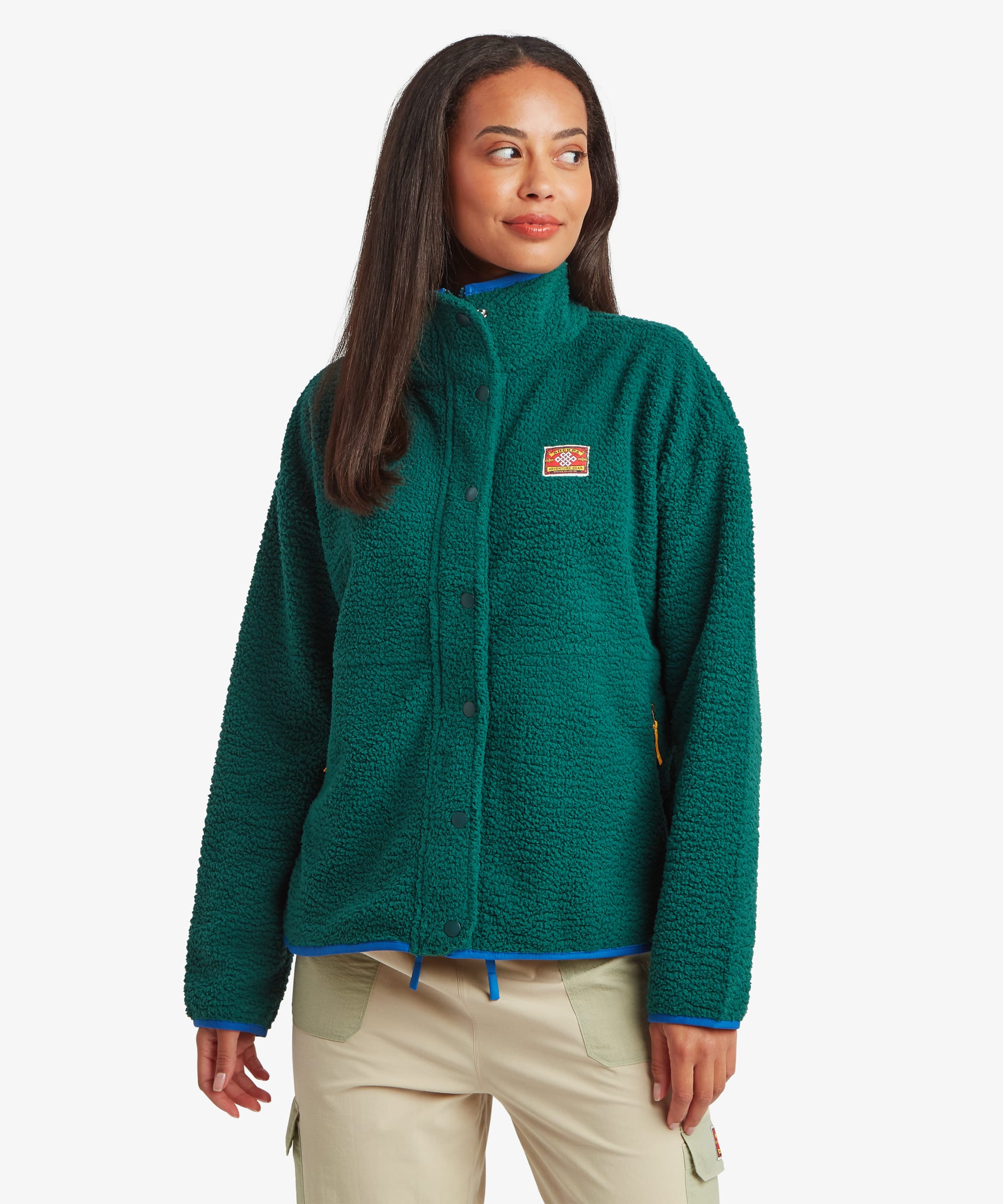 Woman standing facing forward against a plain studio background wearing a deep green full-zip fleece jacket with a high collar and contrasting trim accents. Paired with light beige trousers, the image highlights the jacket's relaxed fit and clean silhouette.