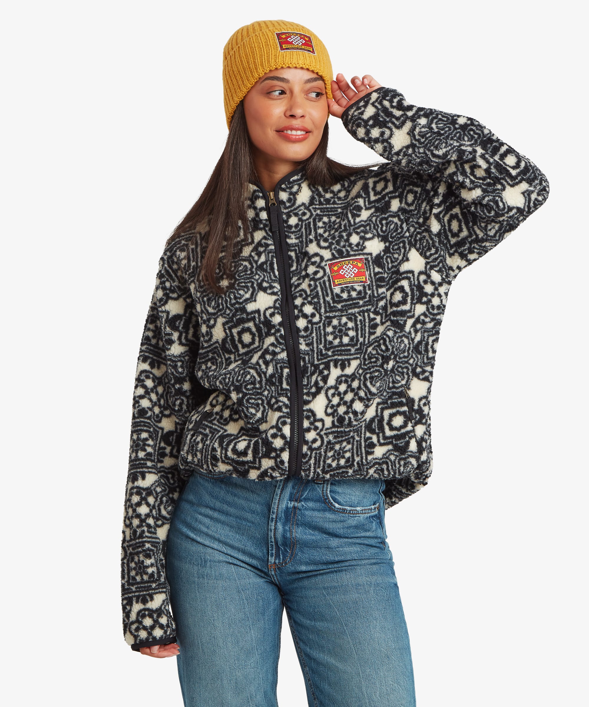 Woman standing against a plain studio background wearing a black full-zip fleece jacket featuring a colourful geometric patchwork-inspired print. She pairs the jacket with blue jeans and a mustard-yellow knit beanie, standing in a relaxed pose that showcases the front of the garment.