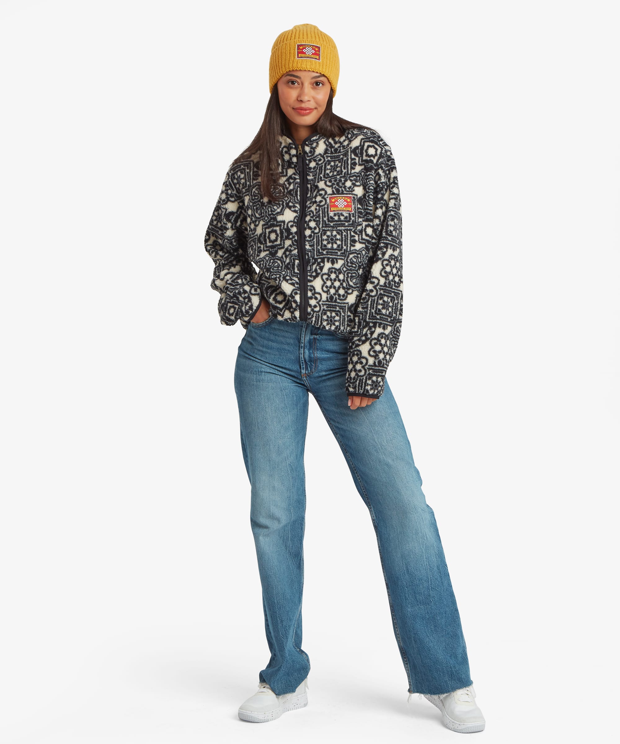 Full-length studio image of a woman wearing a black fleece jacket with a vibrant multicoloured geometric print throughout. Styled with blue jeans and a mustard-yellow beanie, the image highlights the jacket's overall fit and pattern placement.