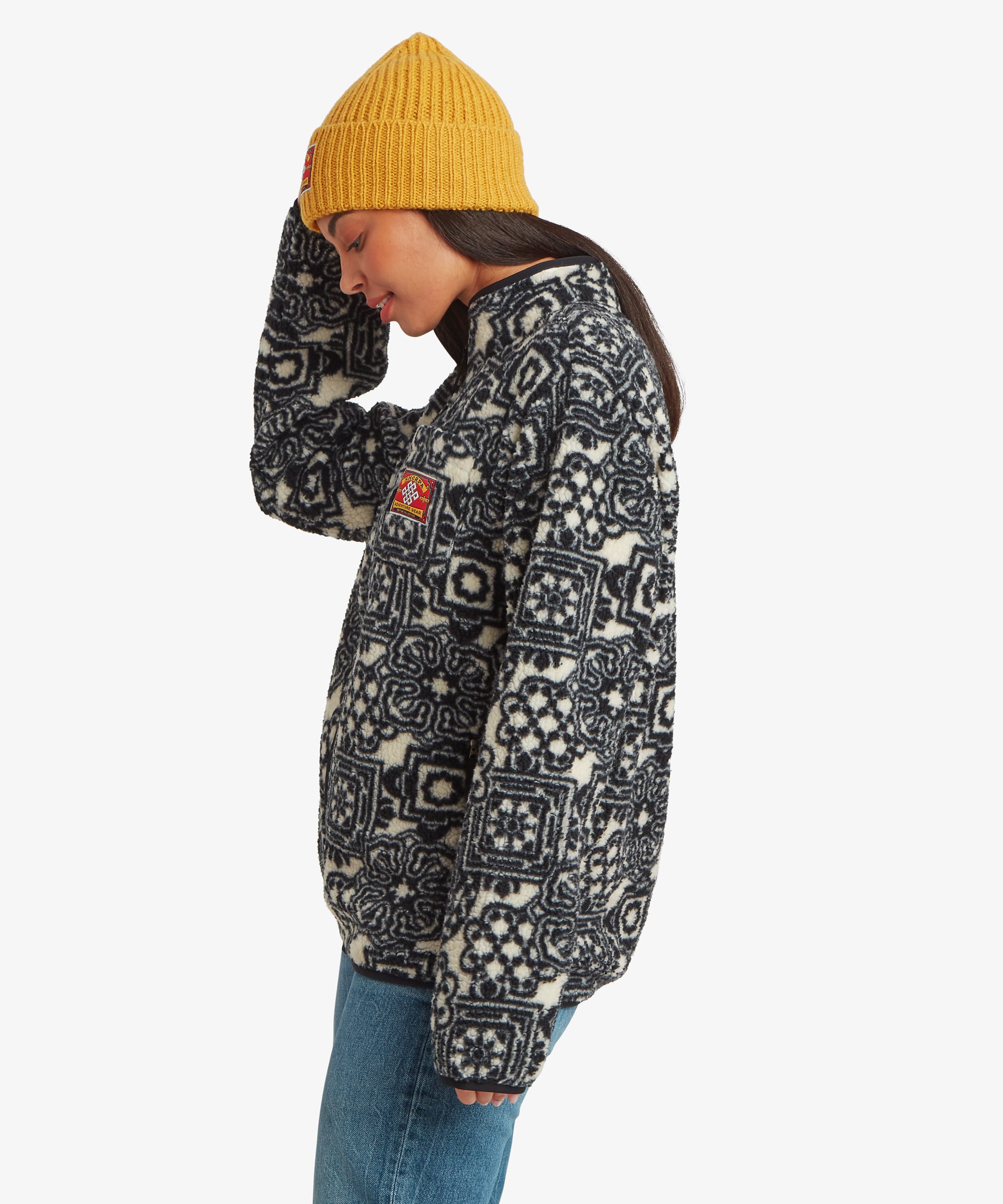 Side-profile studio view of a woman wearing a black fleece jacket covered in colourful geometric motifs. Paired with blue jeans and a mustard-yellow beanie, the image showcases the garment's side profile and relaxed silhouette.
