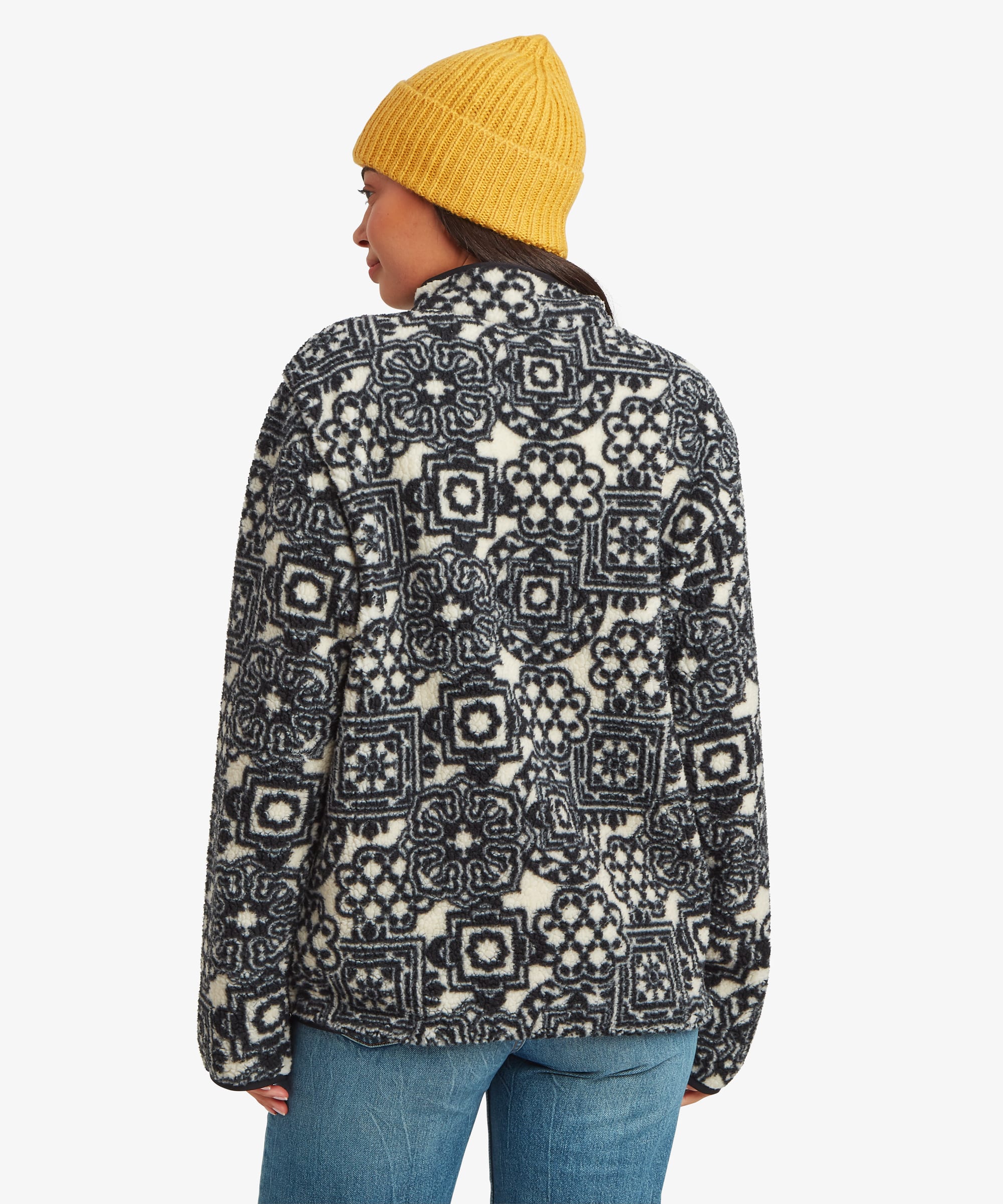 Rear studio view of a woman wearing a black fleece jacket with an all-over multicoloured geometric print. Styled with blue jeans and a mustard-yellow knit beanie, the image displays the pattern coverage across the back and sleeves.