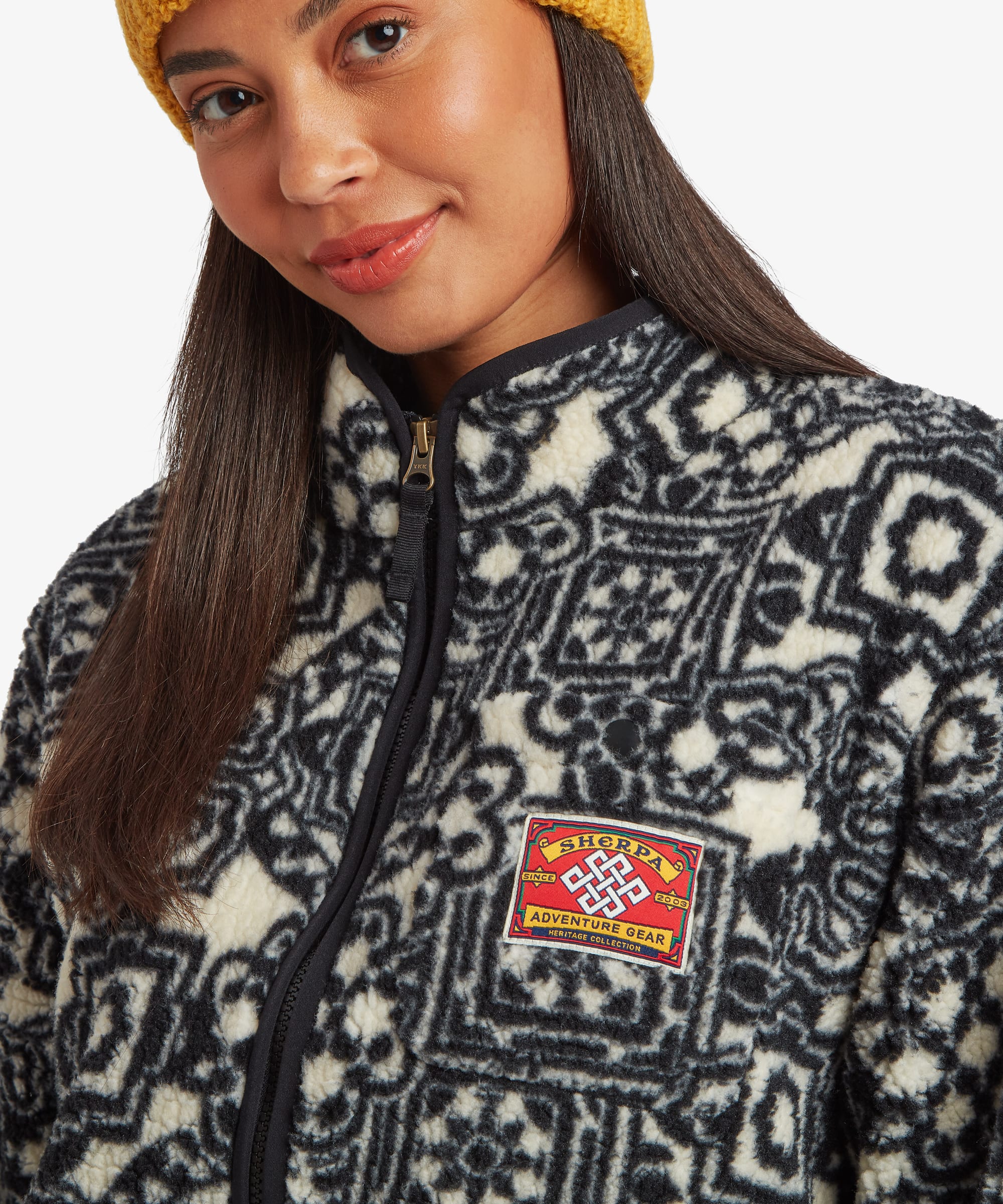 A close-up of a woman wearing a black and cream patterned fleece jacket with a black zipper and a red and yellow 'Sherpa Adventure Gear' logo patch on the chest, paired with a yellow knit beanie against a plain white background.