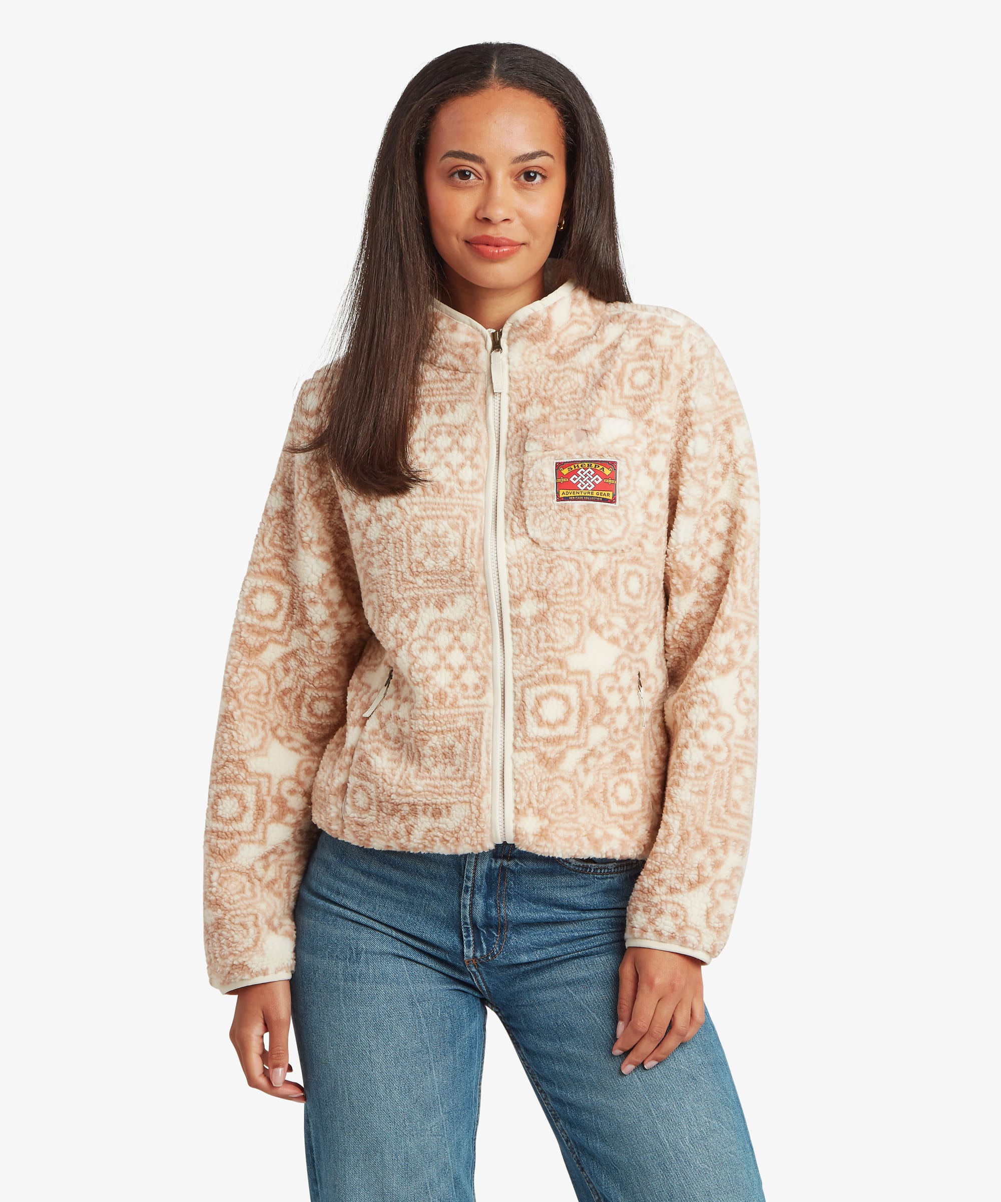 Woman standing facing forward against a plain studio background wearing a cream fleece jacket with an all-over tonal geometric print. Paired with blue jeans, the image clearly displays the jacket's relaxed fit, full zip closure, and high collar.