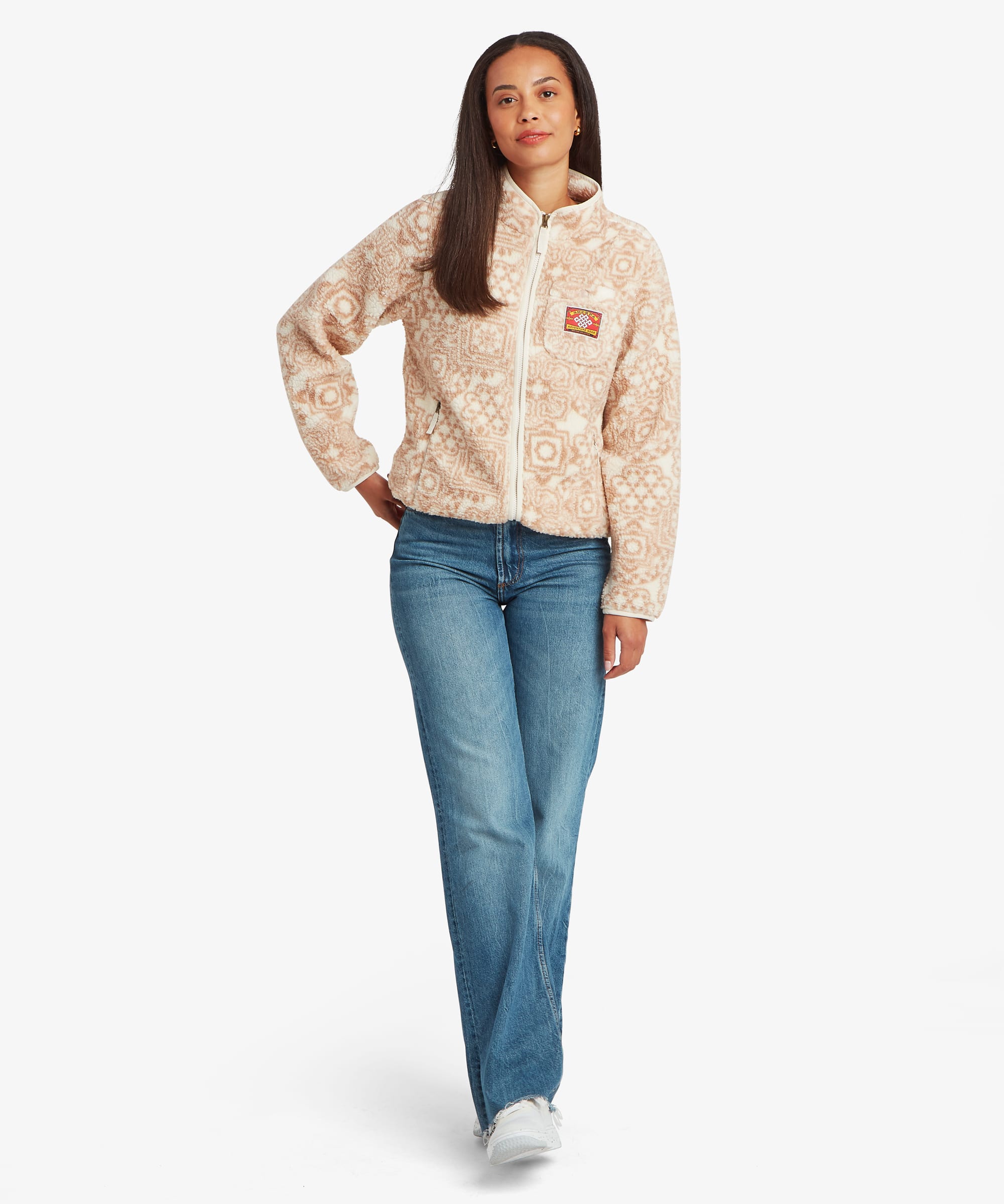 Full-length studio image of a woman modelling a cream printed fleece jacket with subtle geometric motifs throughout. She stands confidently with one hand on her hip, paired with blue jeans and white trainers.