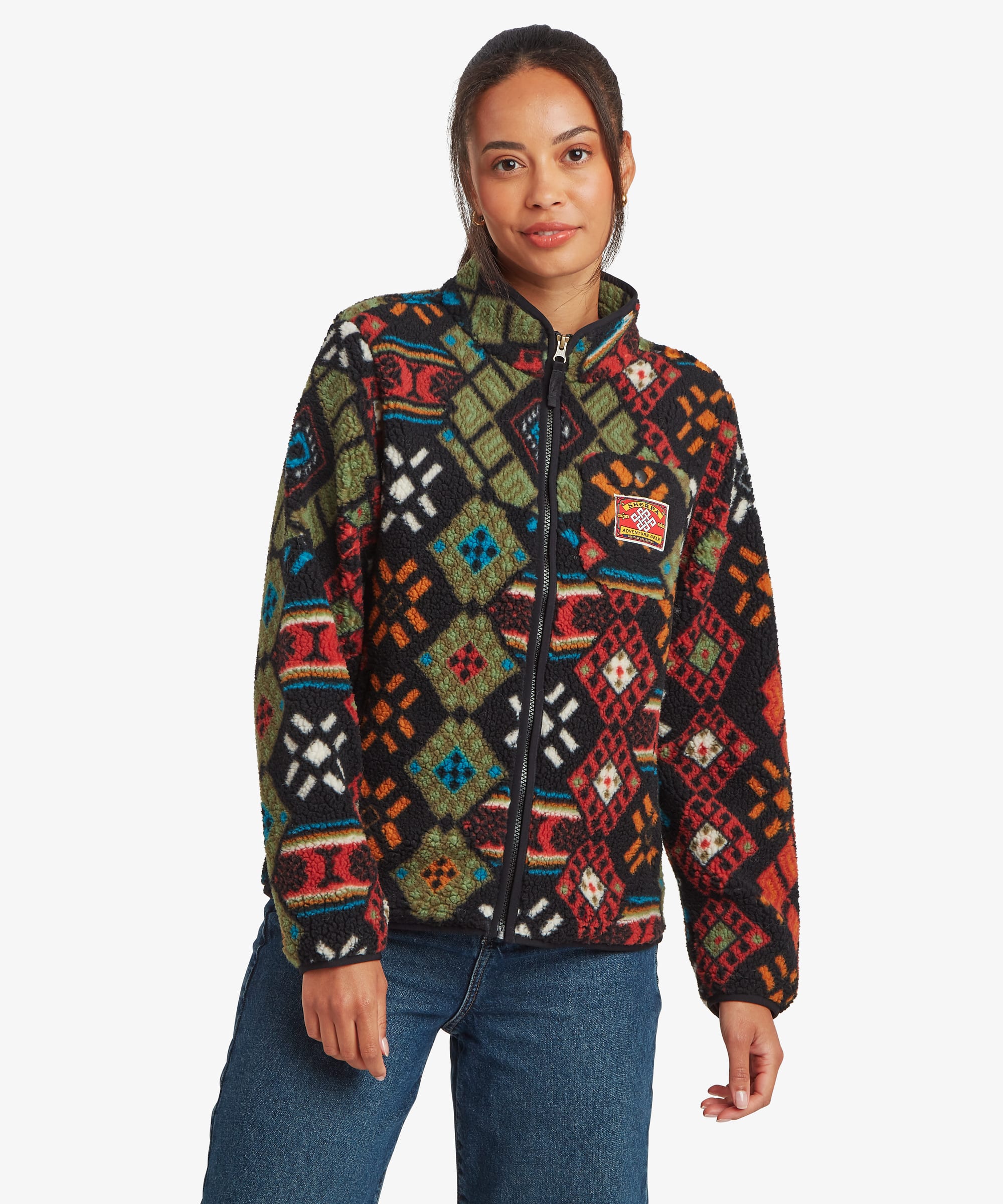 Woman standing facing forward against a plain studio background, wearing a black full-zip fleece jacket featuring a colourful geometric patchwork-inspired print in red, blue, green, and orange tones. The jacket is paired with blue jeans, with her relaxed stance showcasing the overall fit and pattern placement.