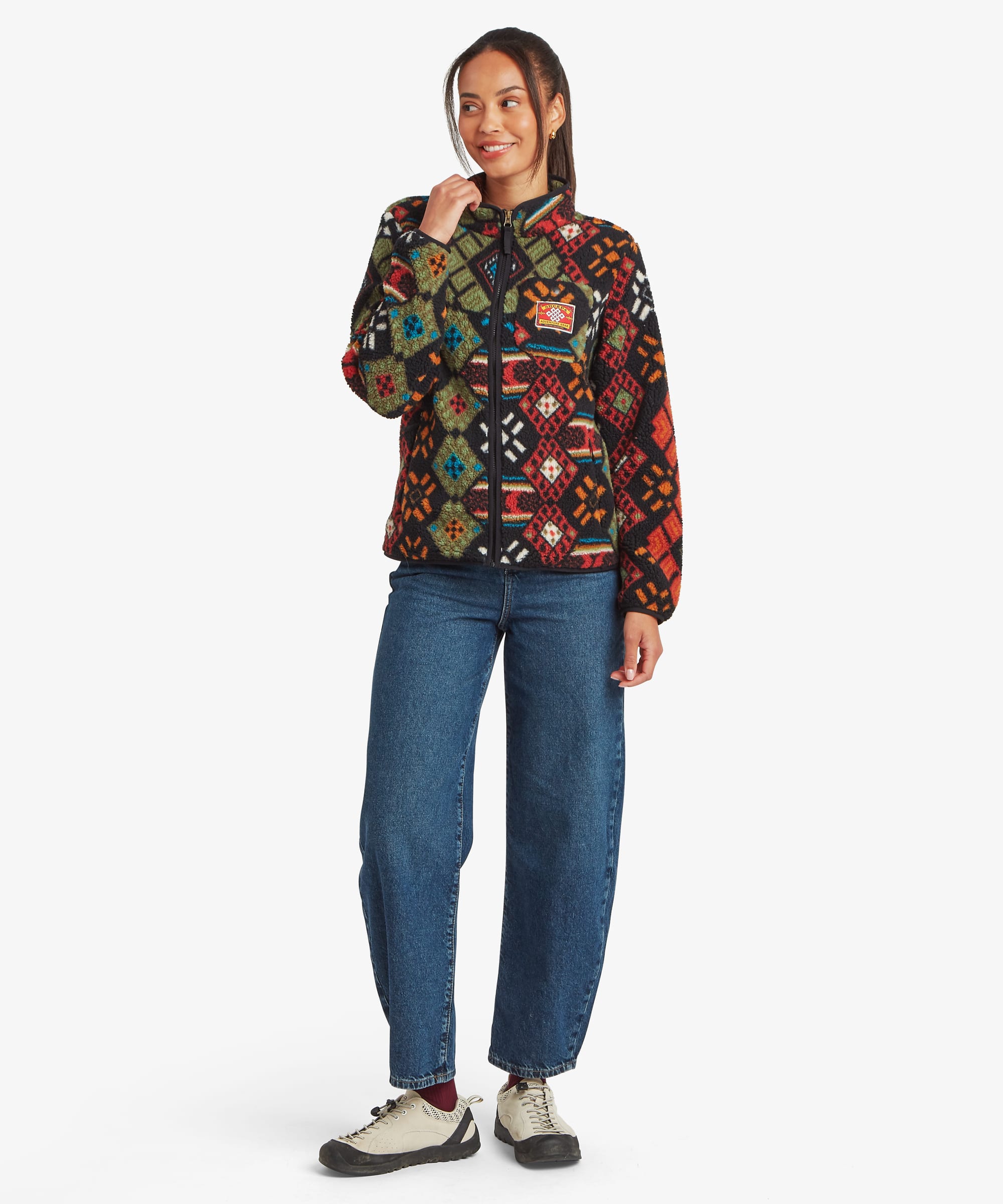 Full-length studio image of a woman wearing a black printed fleece jacket with a vibrant multicoloured geometric design. She stands casually with one hand near her face, paired with blue jeans and trainers, highlighting the jacket's relaxed silhouette.