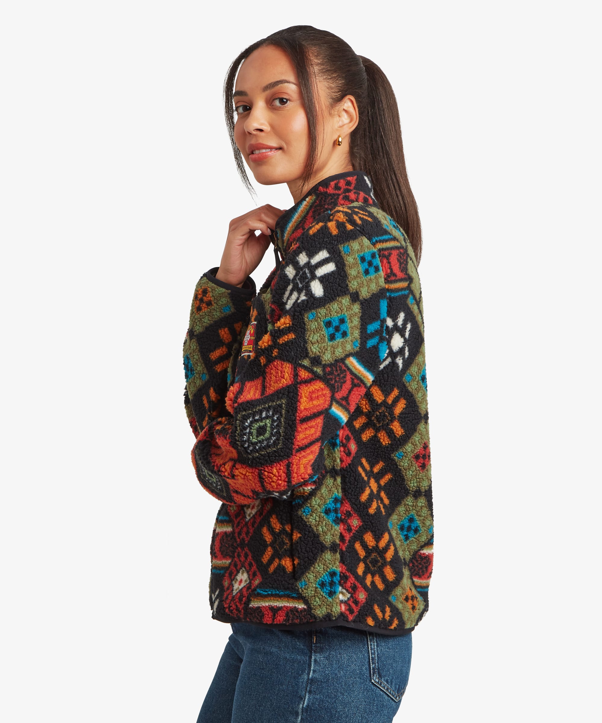 Side-profile studio view of a woman wearing a black full-zip fleece jacket covered in colourful geometric motifs. The image highlights the jacket's side fit, high collar, and relaxed shape when worn with blue jeans.