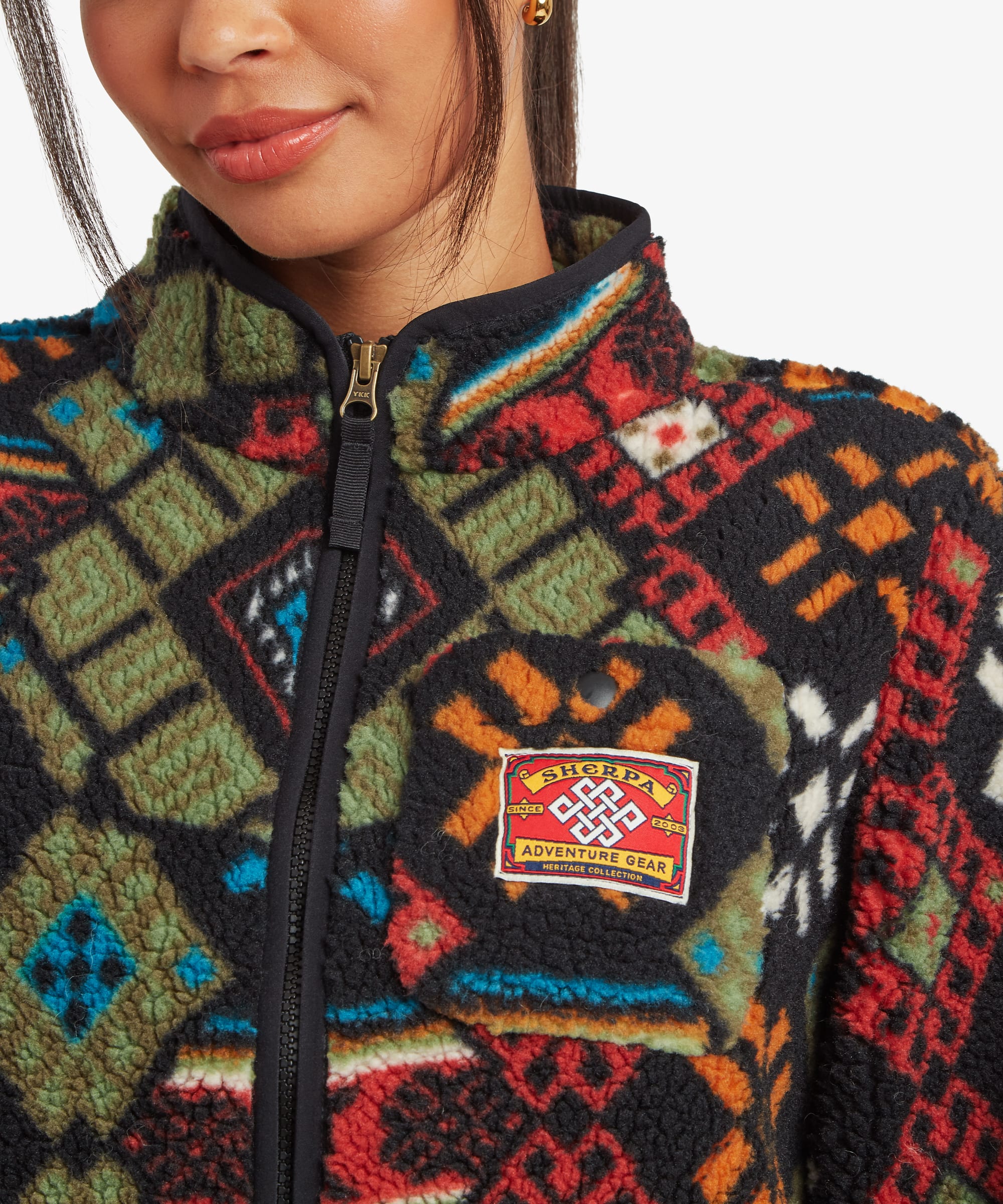 Close-up portrait of a woman wearing a black fleece jacket with a colourful geometric print and stand collar. The cropped image highlights the jacket's zip closure, chest patch detail, and intricate pattern.