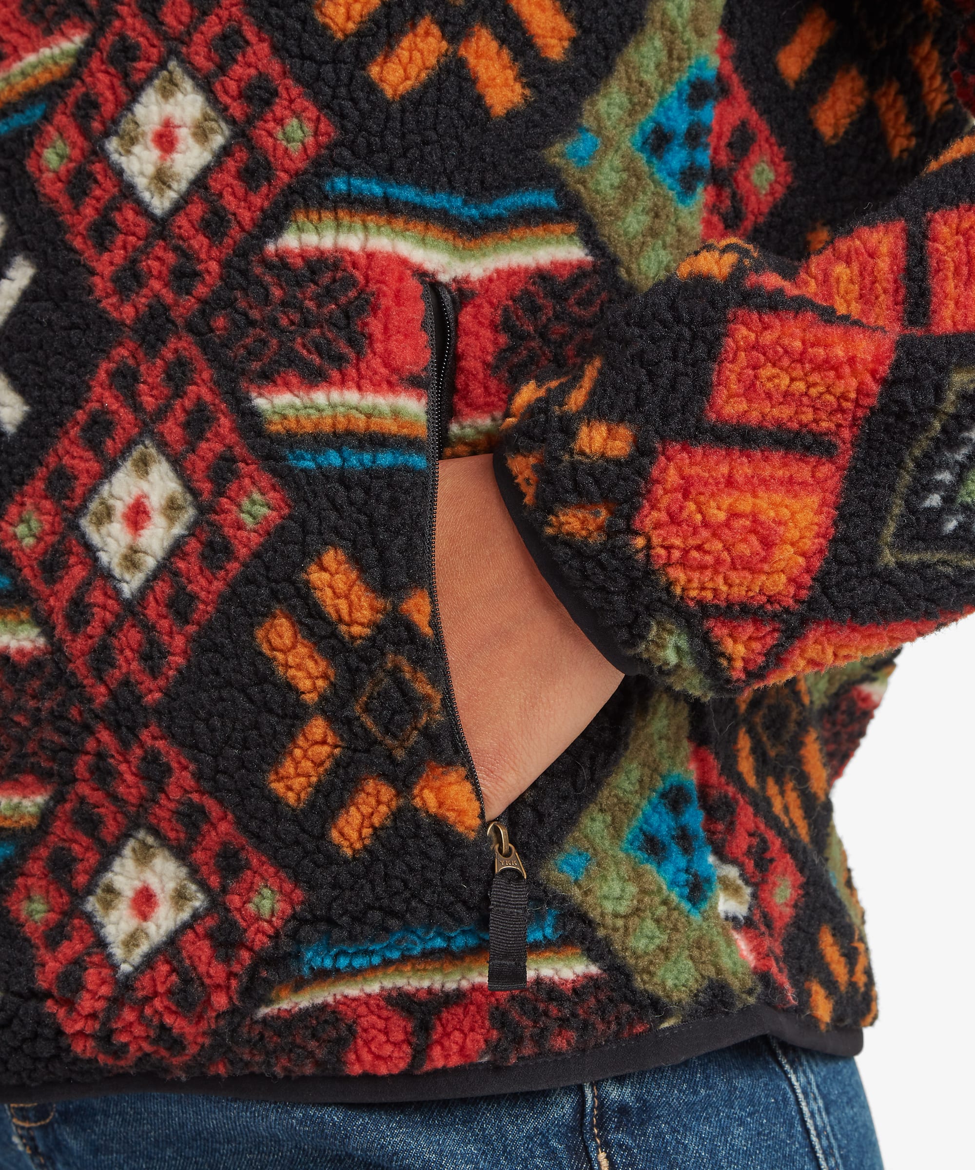 Close-up detail image of the black printed fleece jacket showing the colourful geometric pattern, zippered hand pocket, and textured fleece fabric. The cropped composition focuses on garment construction and print details.