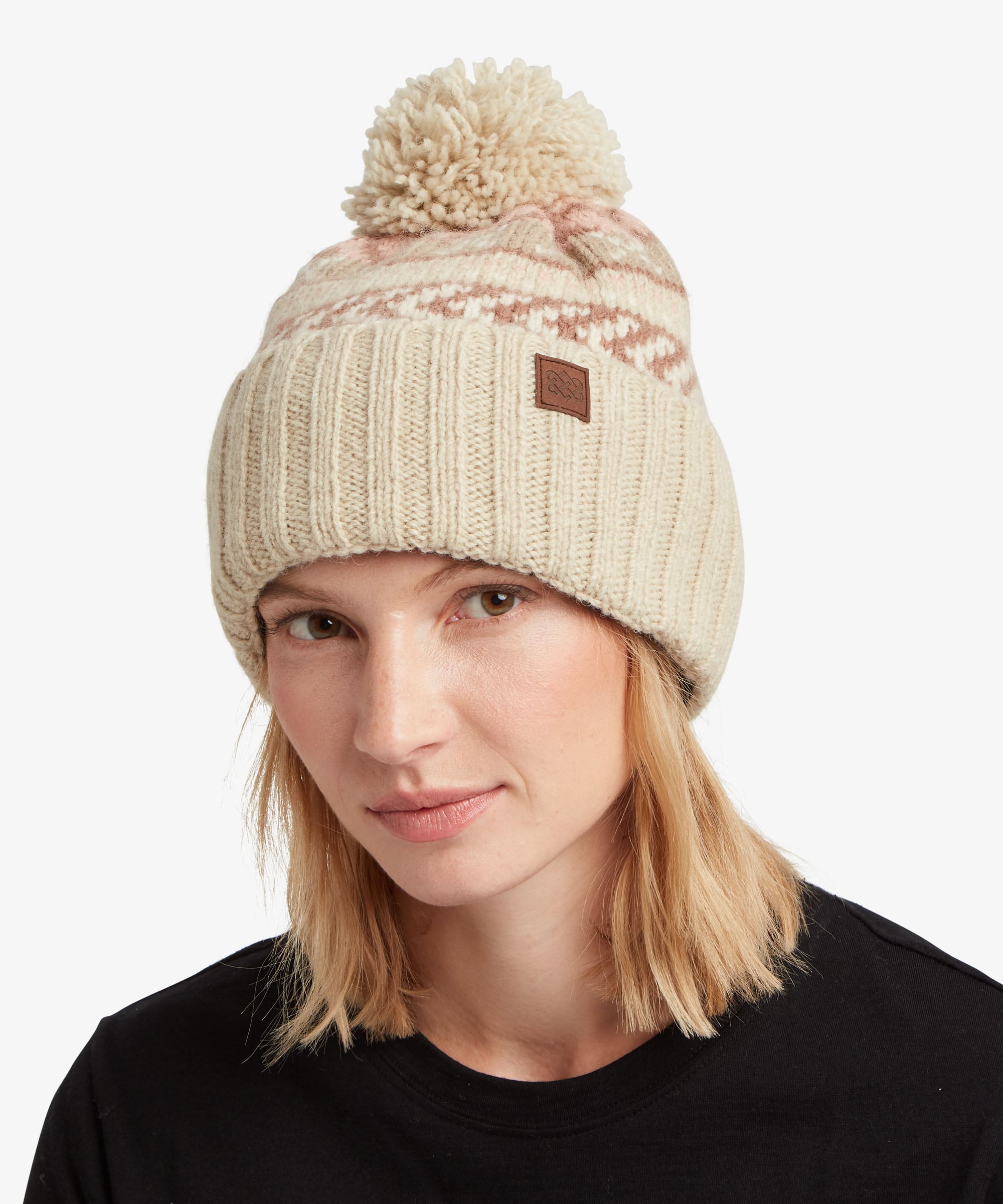 A woman wearing a beige and pink knit beanie with a brown pom-pom and a small brown brand patch.