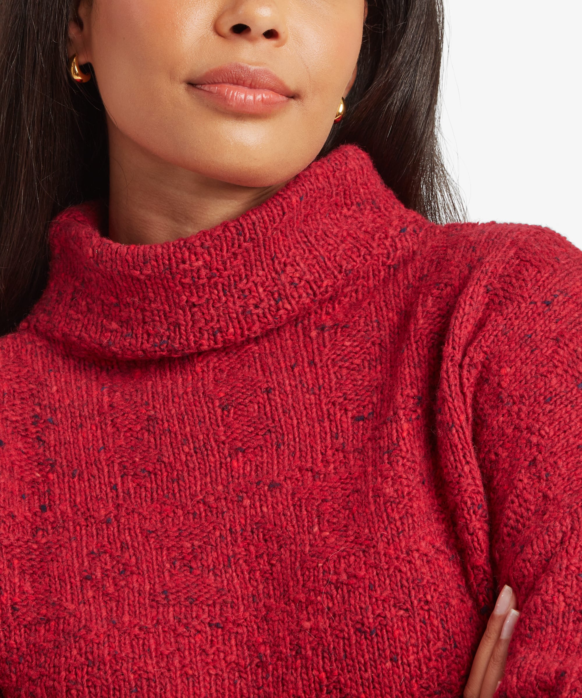 Close-up of a person wearing a chunky knit turtleneck sweater in heathered red with subtle dark flecks, paired with small gold hoop earrings against a plain light background.