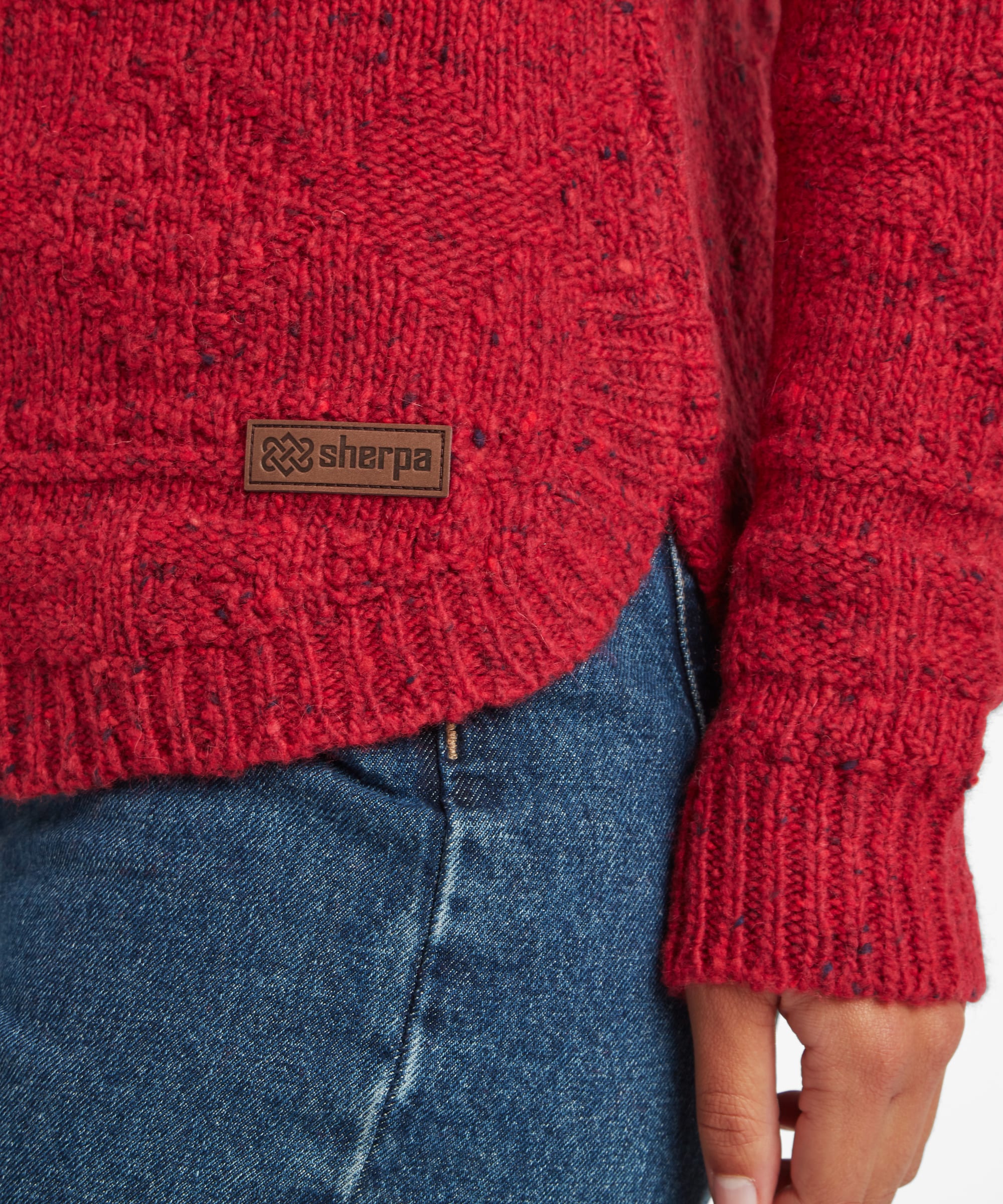 Close-up of a woman wearing a red textured knit sweater with a brown Sherpa brand patch on the chest, paired with blue jeans.